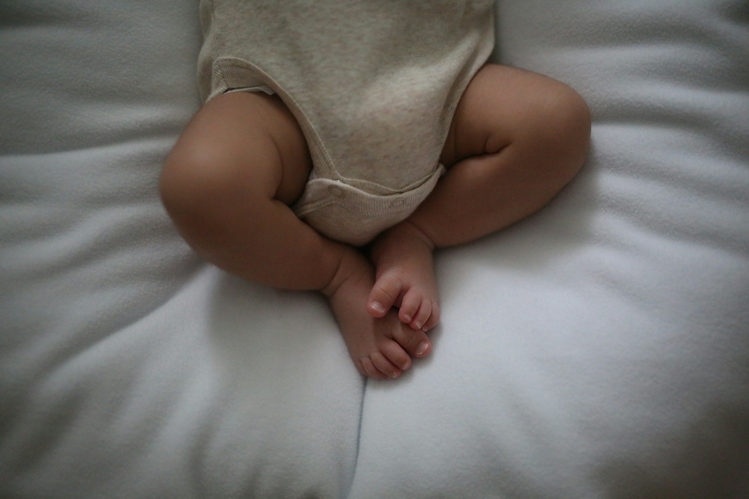 Close-up of a baby's legs and feet, sitting on a white surface, wearing a beige onesie.