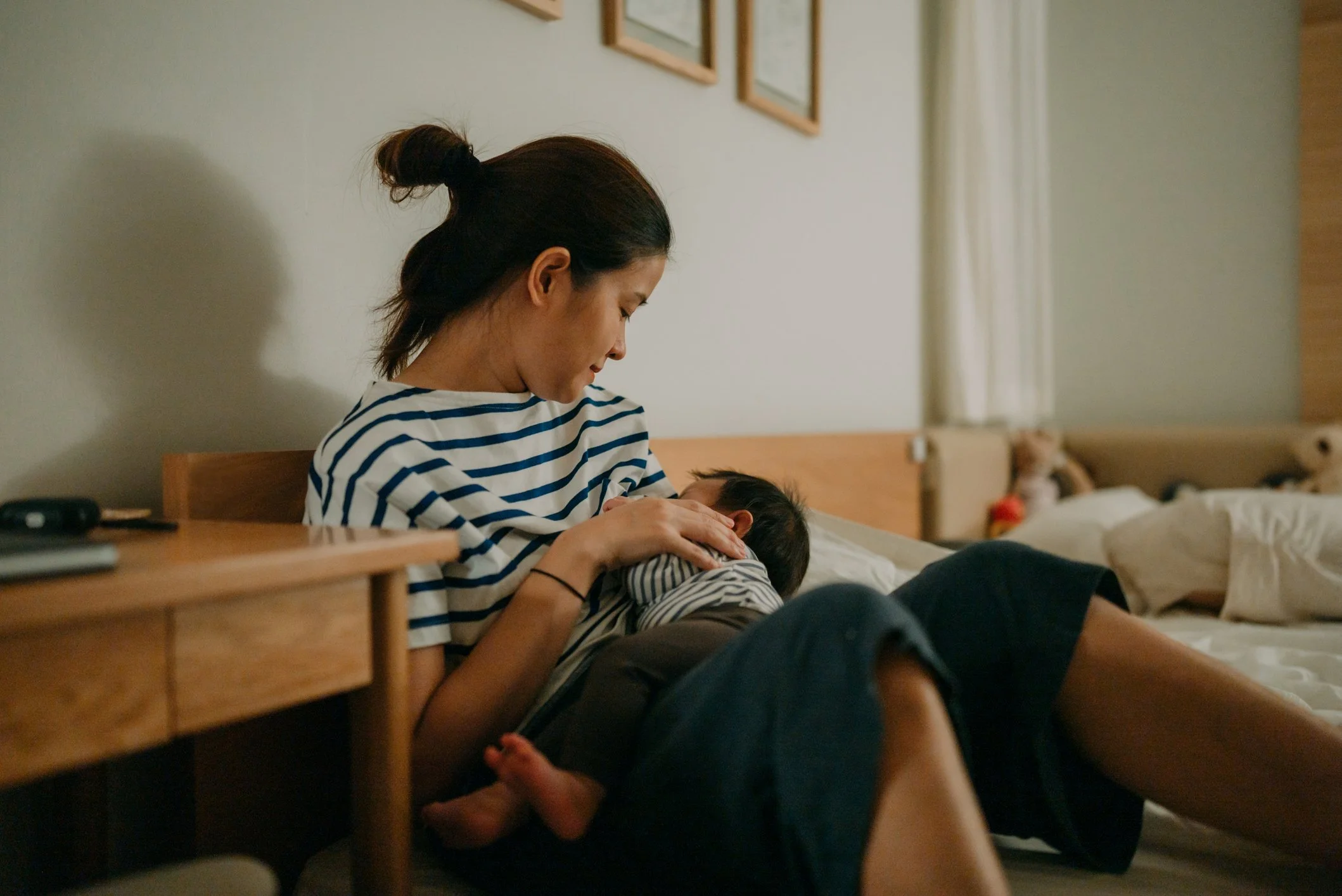 A woman with dark hair tied in a bun wearing a striped shirt is sitting on a bed, holding and comforting a small child in striped pajamas. The scene appears to take place in a bedroom, with a nightstand and framed pictures on the wall visible in the background.