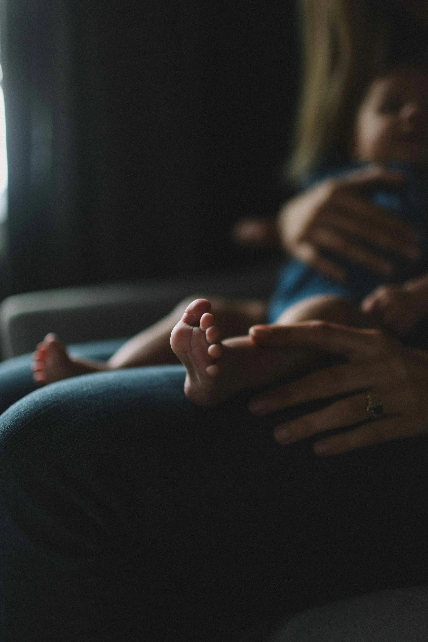 A young child sitting on a person's lap, holding a phone and looking at it, with another person's hand gently supporting the child's waist. The background is dark and the photo is softly lit.