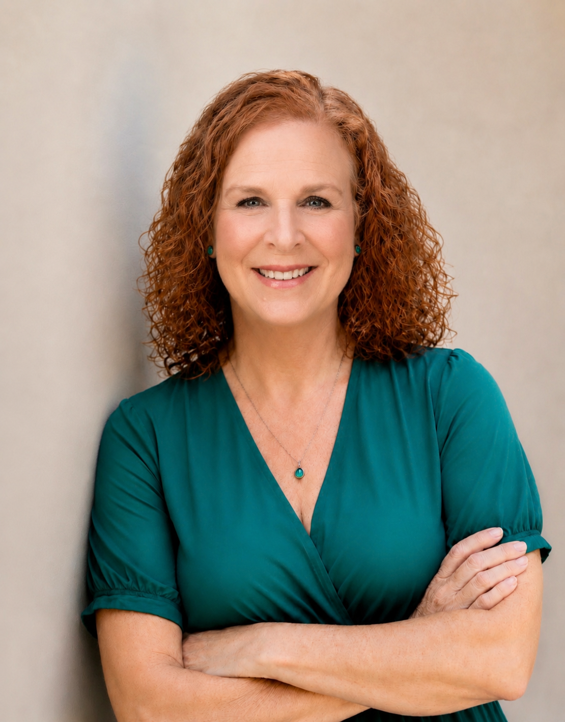 A smiling woman with curly red hair, wearing a teal dress and jewelry, posing with arms crossed against a plain beige background.