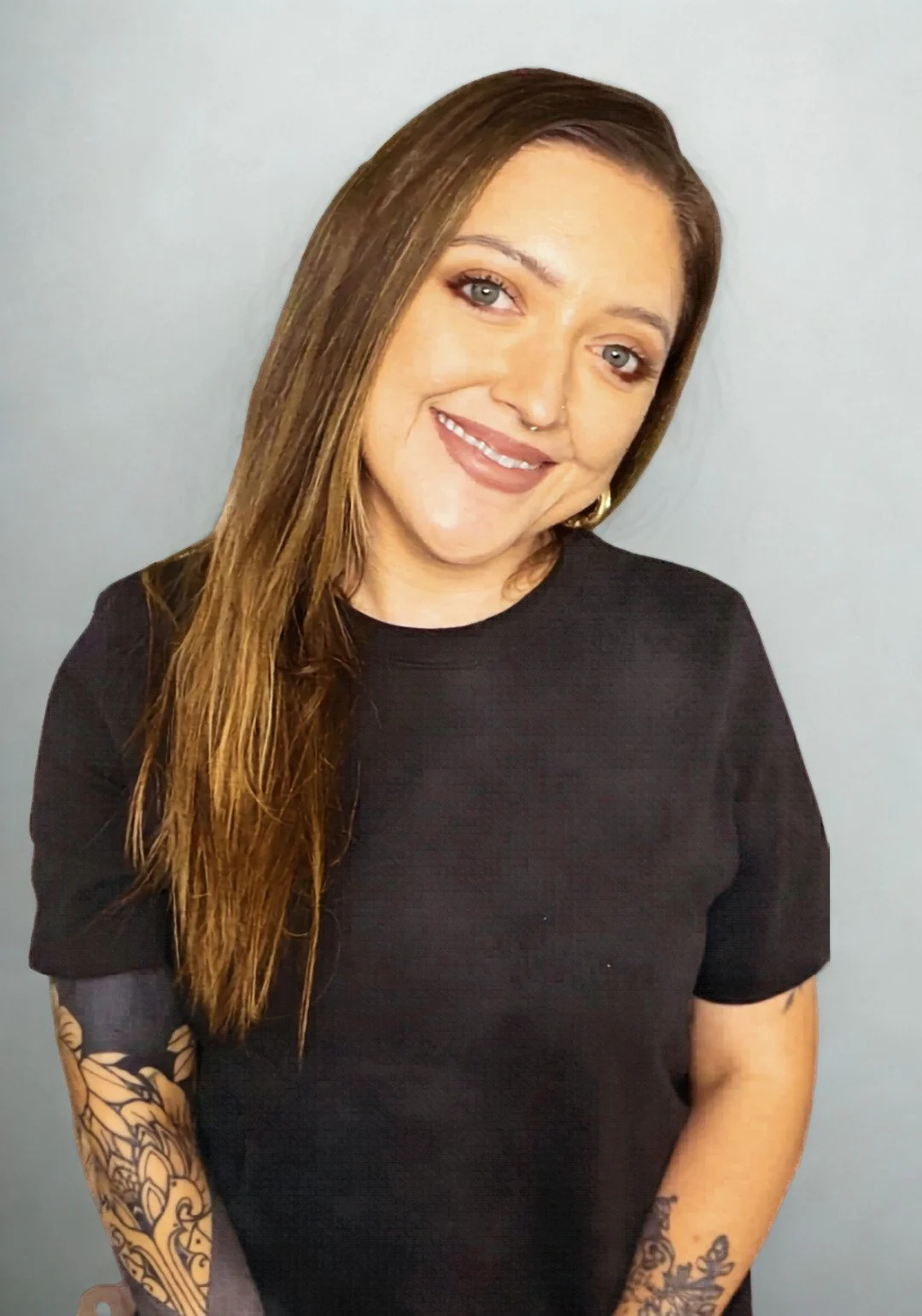 A postpartum doula with long brown hair, blue eyes, and tattoos on her arms, smiling and wearing a black shirt, standing against a light gray background in Sacramento California