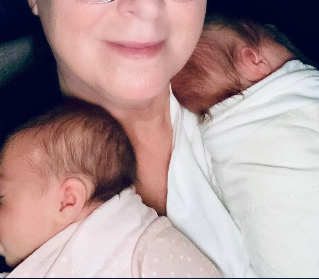 A woman with glasses and short blonde hair smiling while holding two sleeping babies with light brown hair, one on each shoulder.