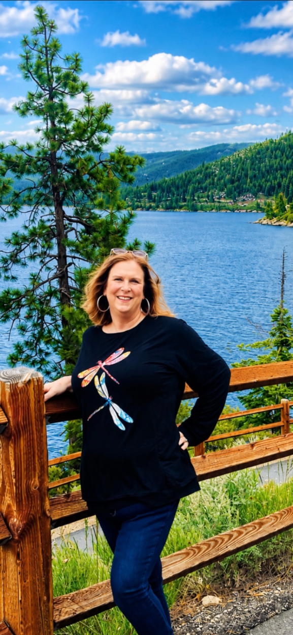 A woman with blonde hair wearing a black shirt with a dragonfly design, standing outdoors near a lake with a wooden railing, surrounded by green trees and hills under a partly cloudy sky.