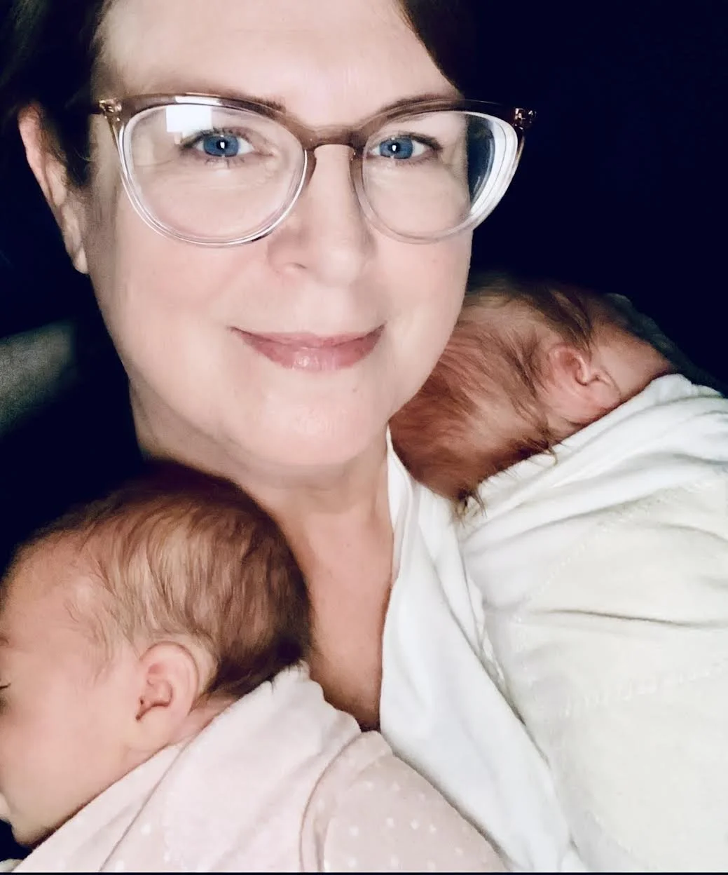 A woman with glasses smiling, holding two sleeping babies with reddish hair close to her chest.
