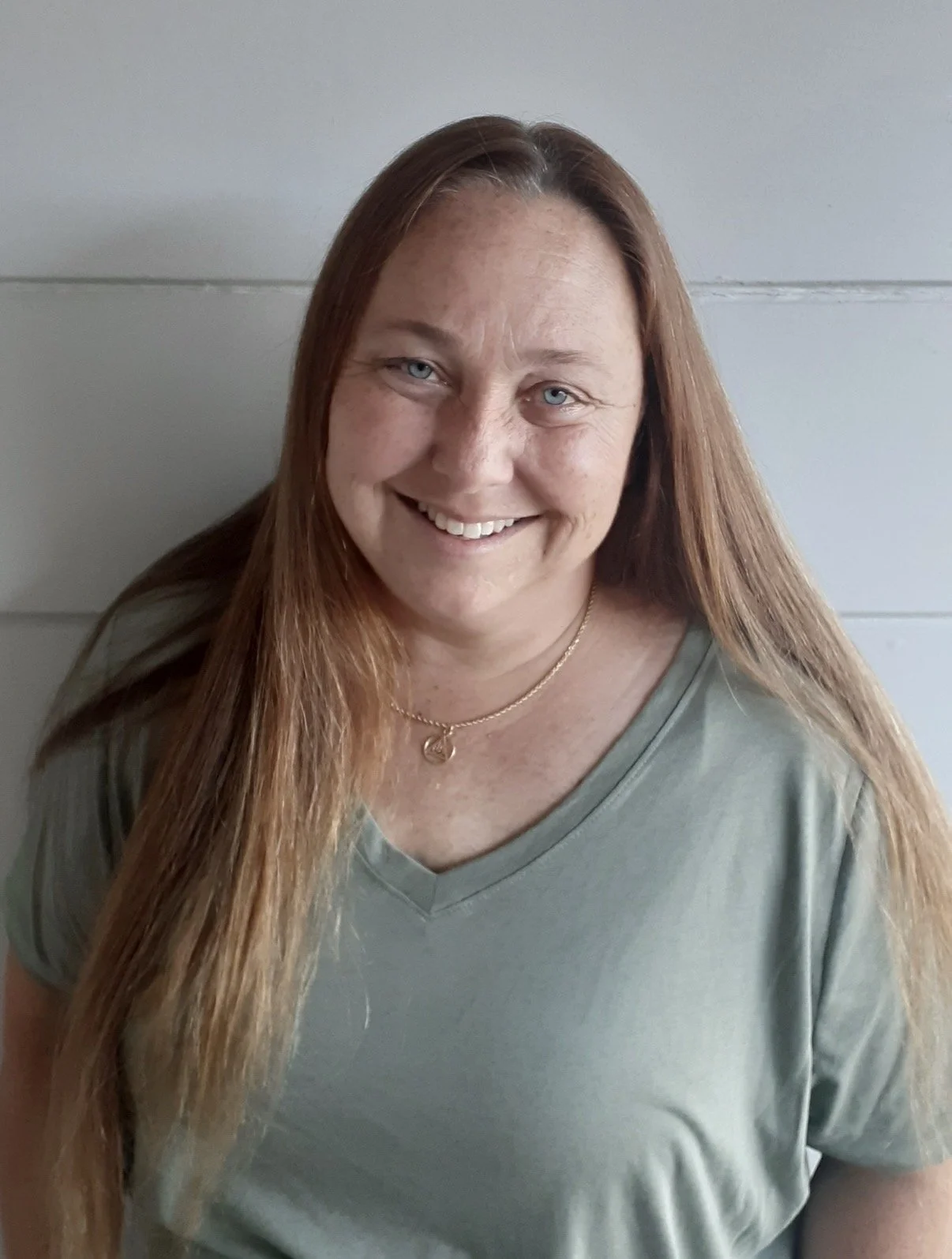 A smiling woman with long red hair, wearing a gray shirt and a gold necklace, standing in front of a light-colored wall.