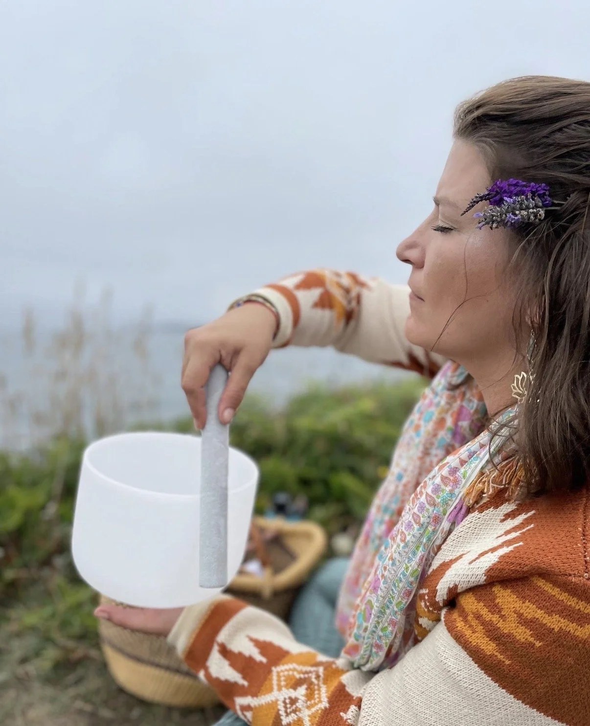 A woman with a lavender accessory in her hair is sitting outdoors by a body of water, holding a white cup with a straw and stirring it with a spoon.