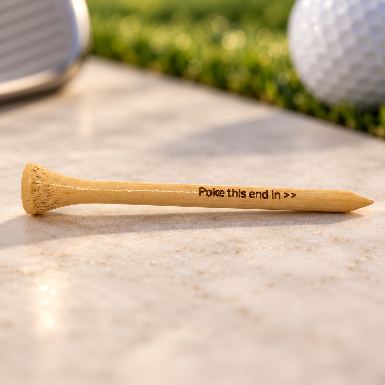 A wooden golf tee resting on a flat surface with inlaid text that reads, 'Poke this end in   '. In the background, there's a golf ball and part of a golf club, with greenery and sunlight.