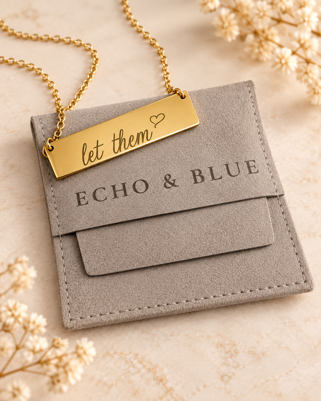 A gold necklace with a rectangular pendant engraved with "let them" and a small heart, resting on a gray suede cardholder with the brand name "ECHO & BLUE". The scene is decorated with white flowers on a beige surface.