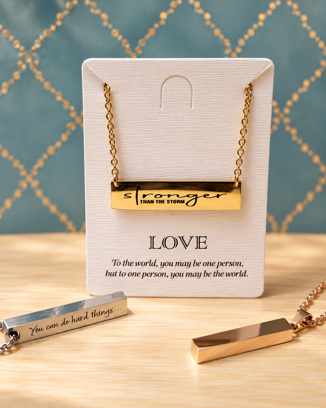 Jewelry display featuring a gold bar necklace with the words 'stronger than the storm' on it, and two matching bracelets with engraved inspirational messages, all on a wood surface against a blue background with a gold chain pattern.
