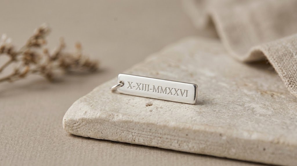 A silver rectangular charm engraved with Roman numerals X-XIII-MMXXVI, resting on a flat stone surface with fabric and dried flowers nearby.