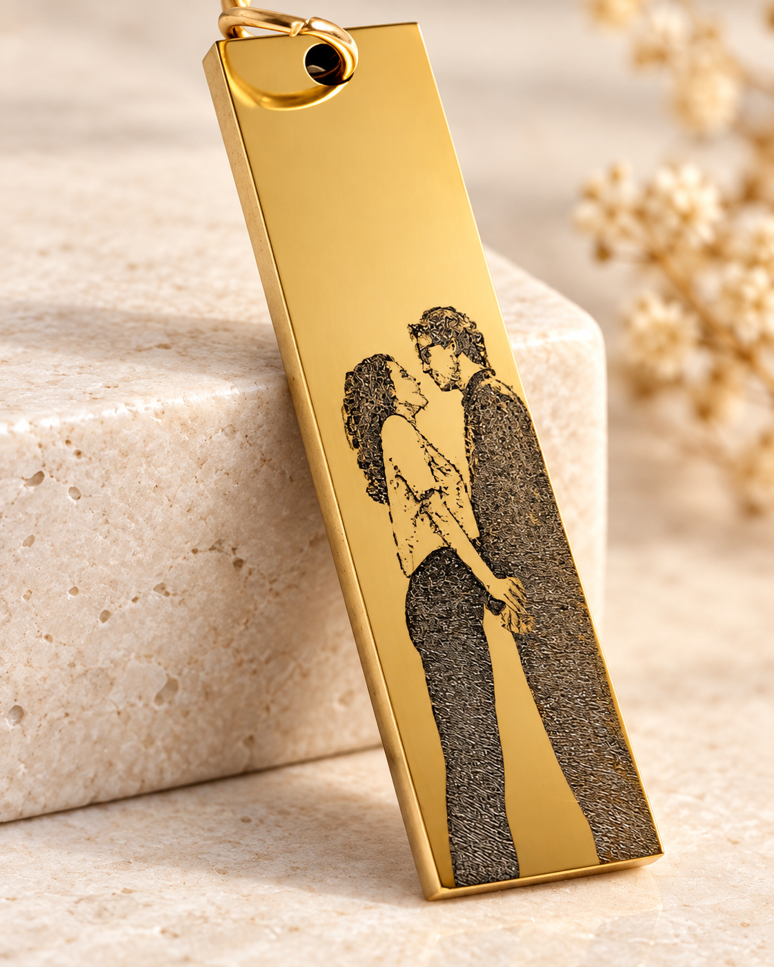 A gold-colored keychain with a black illustration of a man and woman facing each other, standing close, on a beige surface with a blurred background of white flowers.