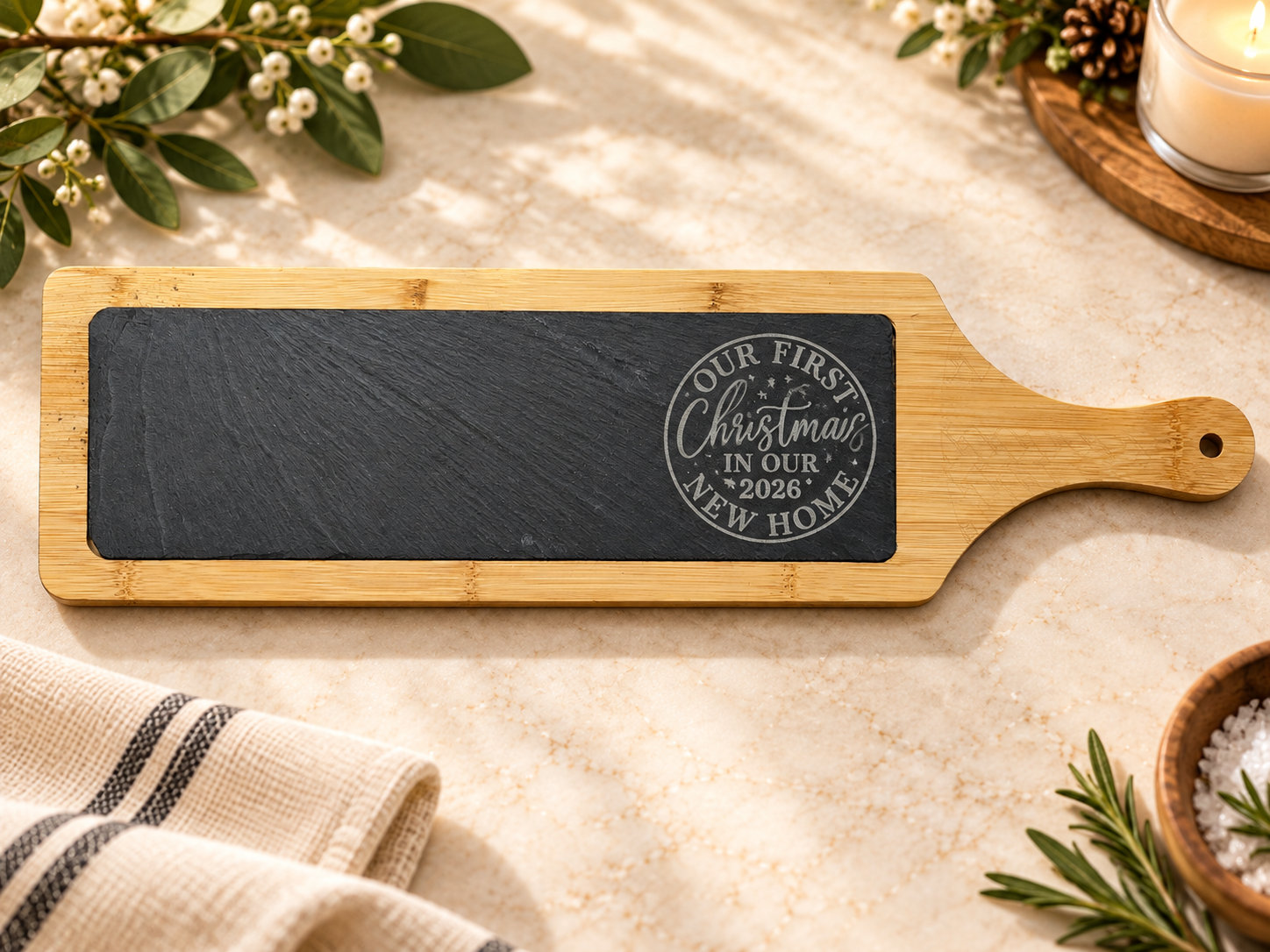 A wooden tray with a black chalkboard surface that has a circular stamp with text and decorative elements, reading 'Our First Christmas in Our New Home 2026'. The tray is on a cream-colored marble surface with holiday decorations including greenery, 