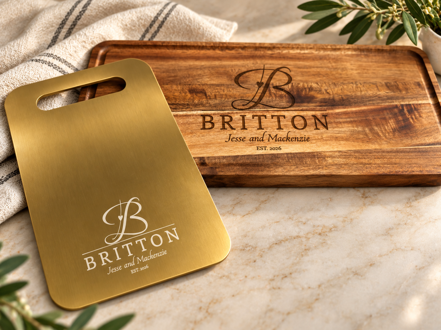 A gold metal serving tray with a monogram, a wooden engraved cutting board with a monogram and text, and some greenery in the corner, on a marble surface.