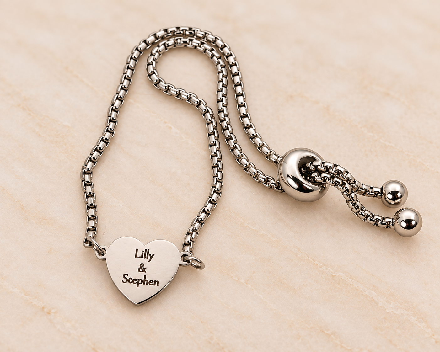 A silver bracelet with a heart-shaped pendant engraved with the names 'Lilly & Scephen' and a sliding clasp with two balls, placed on a light-colored surface.