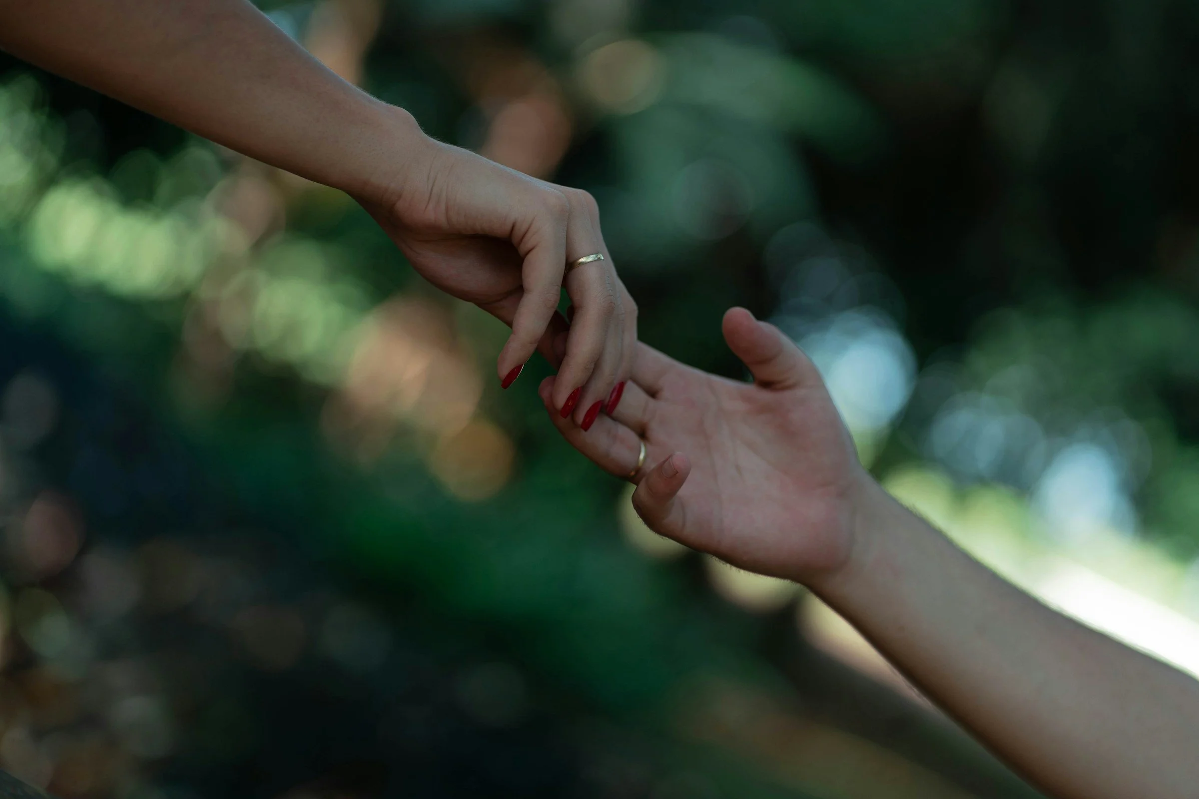 A person's hand with a wedding ring is being held by another person's hand in an outdoor setting with a blurred, green leafy background.