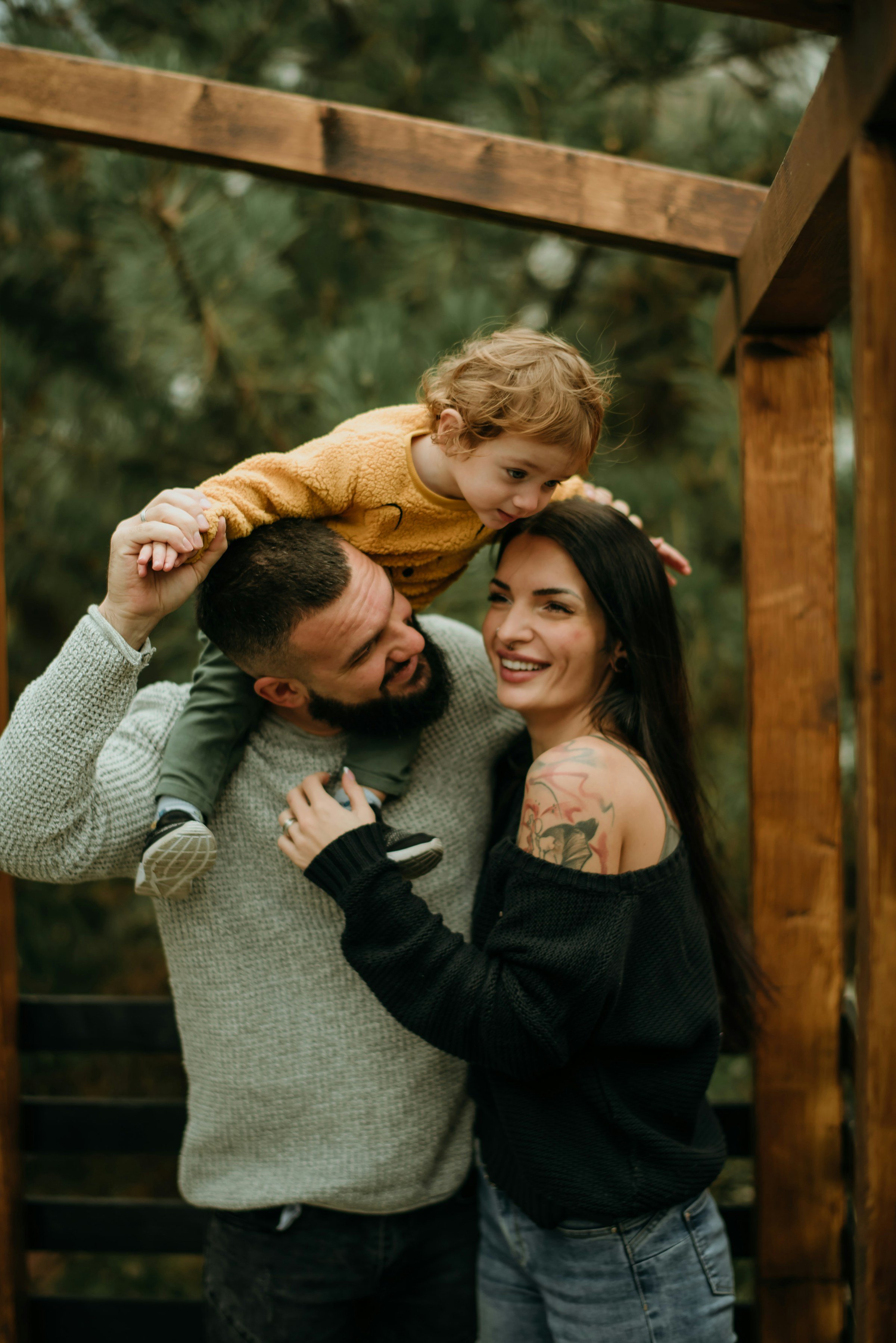 A family of three with a man, woman, and child playing outdoors among trees. The man lifts the child above his head, and the woman smiles, embracing the man. The child is wearing a yellow sweater. The scene is warm and joyful, with greenery in the background.