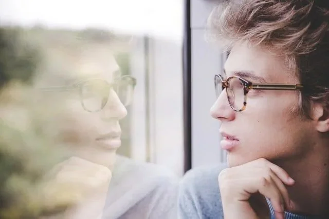 A young man with glasses looking out a window, his reflection visible on the glass.