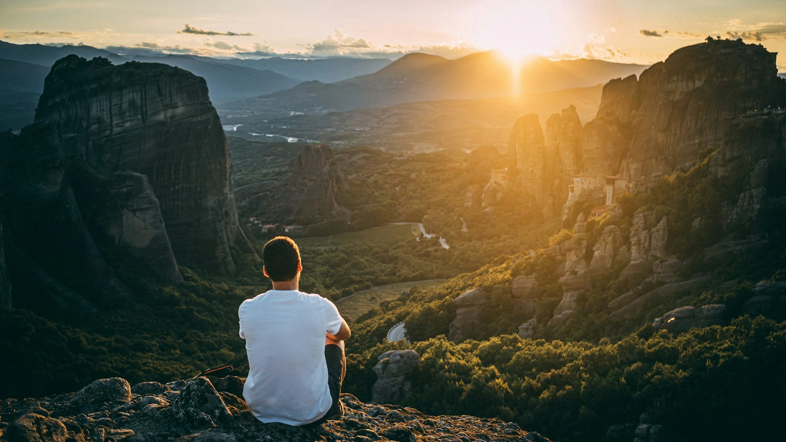 A person sitting on rocky terrain, facing away, watching a sunset over a mountainous landscape with rock formations and green valleys.