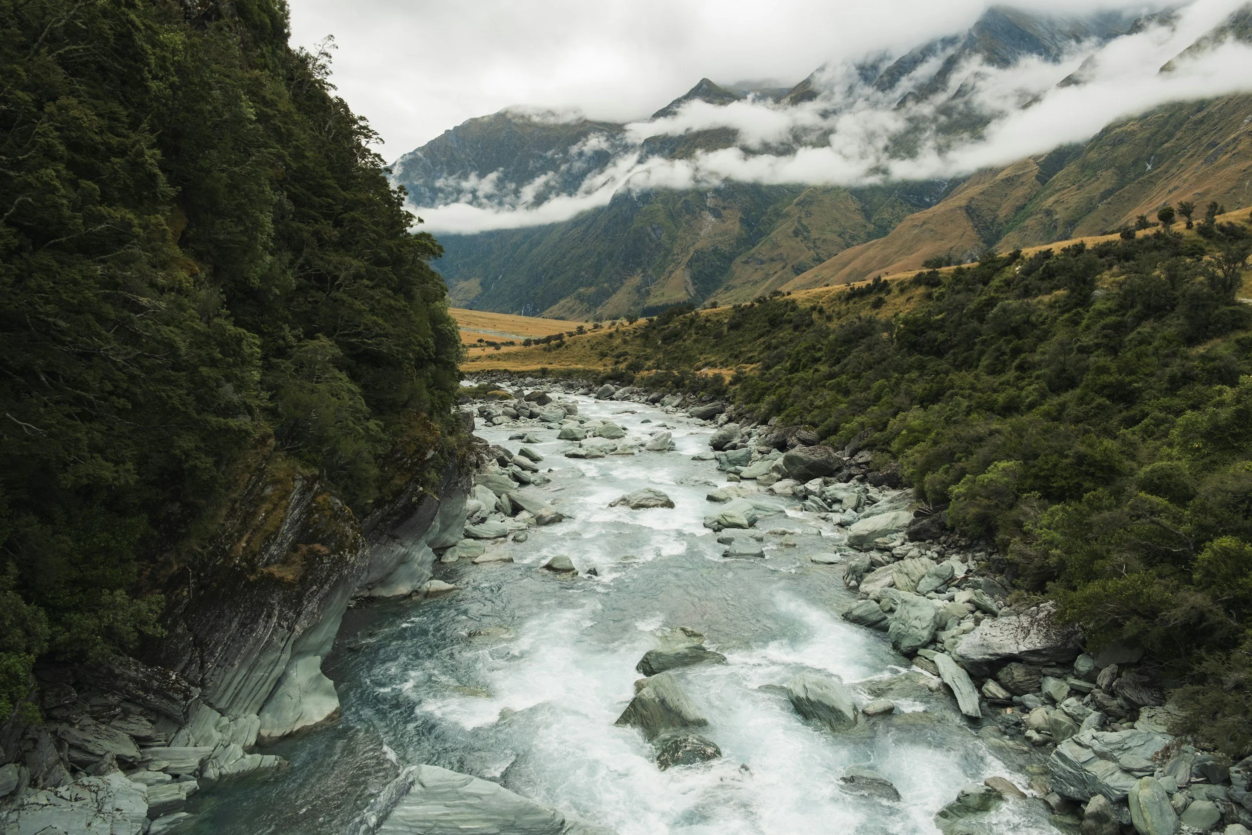 A river flowing through a lush, green valley with mountains in the background partly covered with clouds.