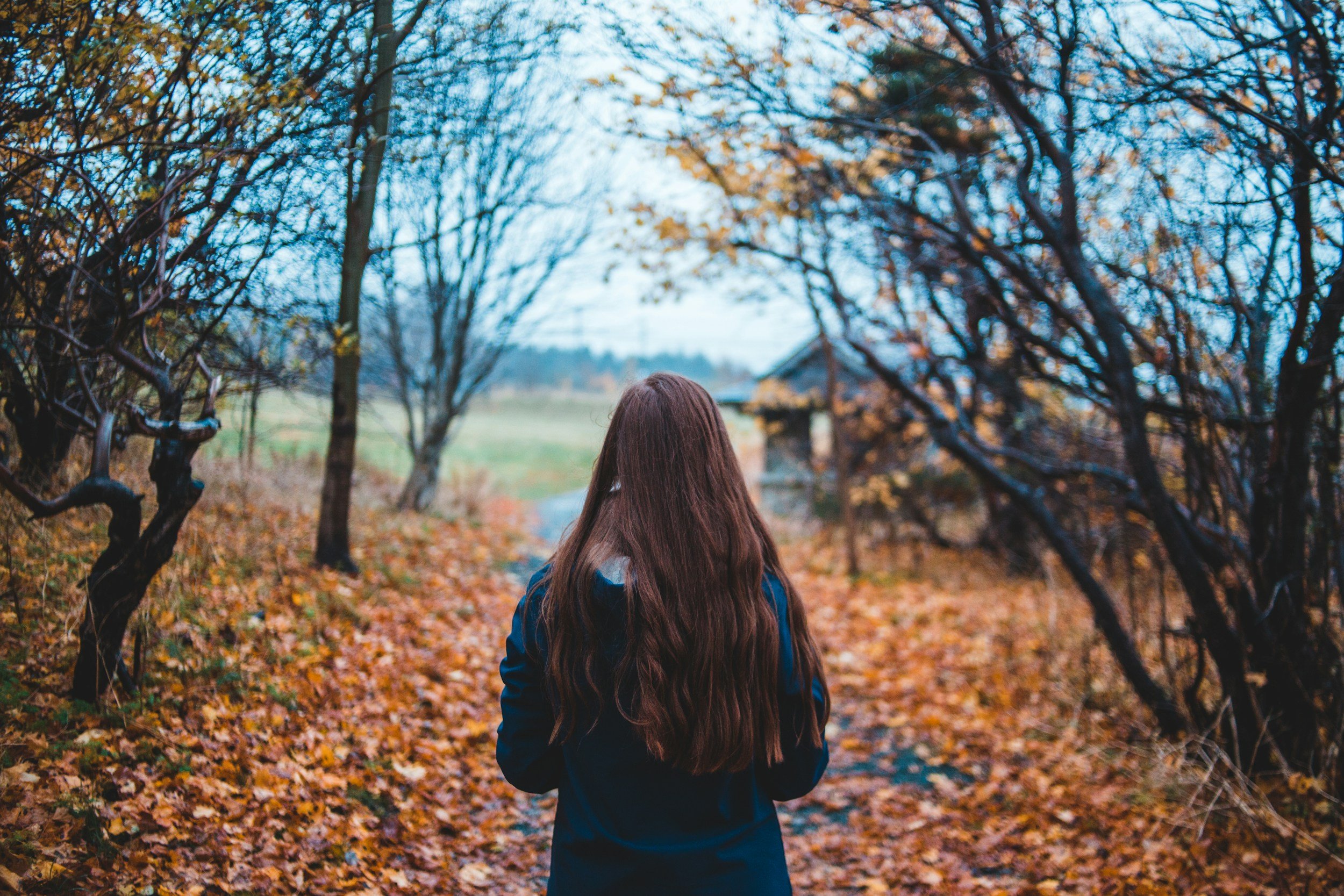 A woman with long brown hair walking through a leaf-covered path in a park with autumn foliage on trees.