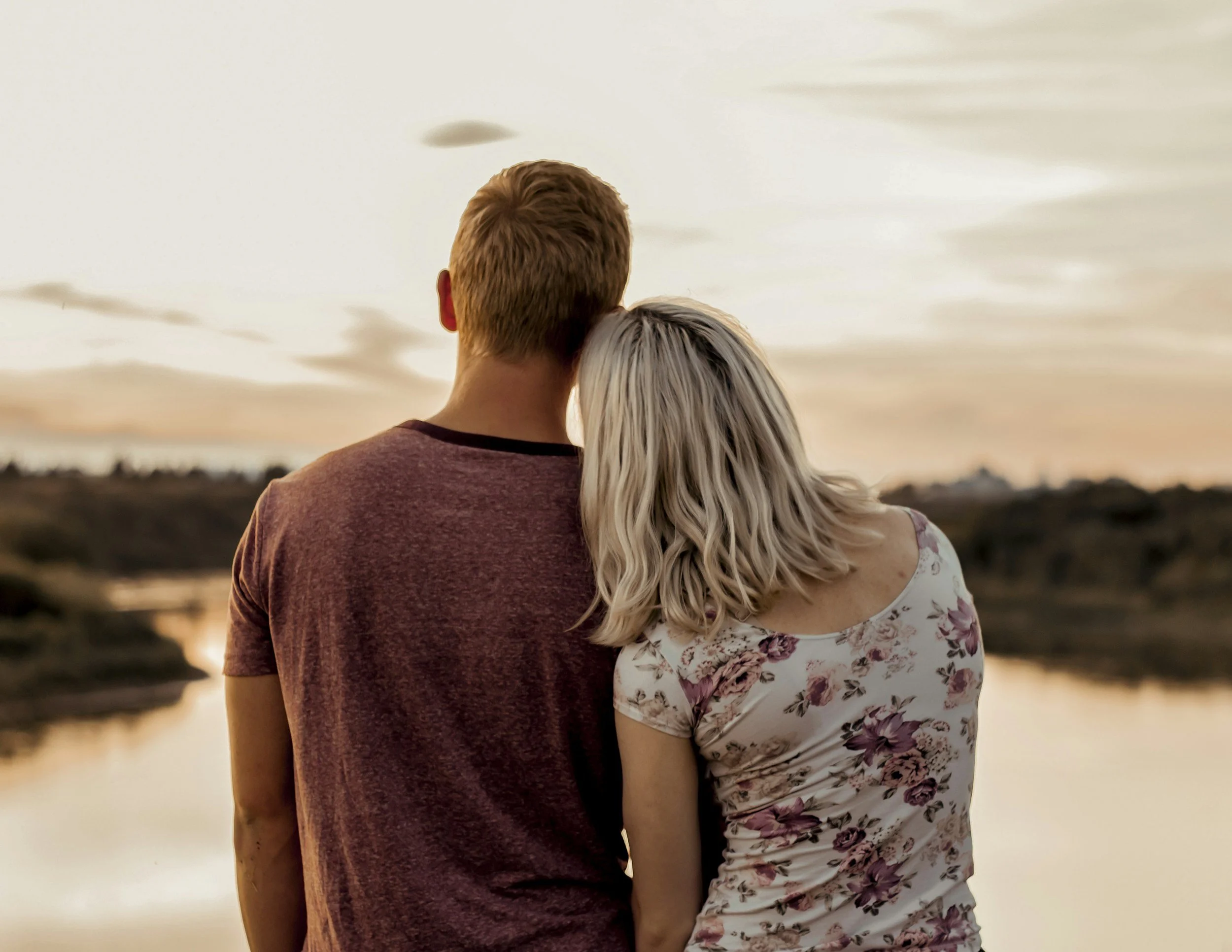 A couple stands close together outdoors during sunset, facing away from the camera, with the sky glowing with warm colors.