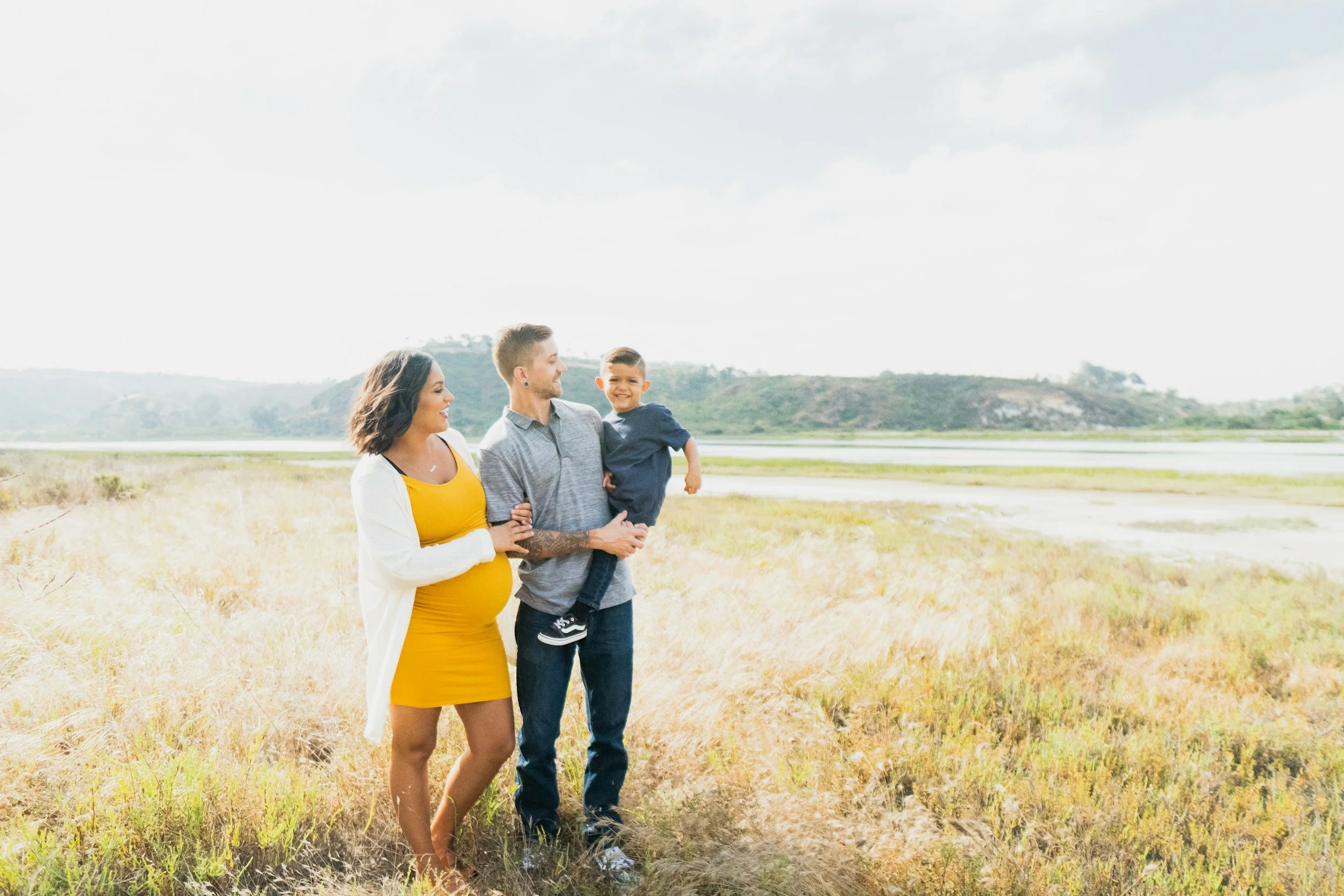 A family of three walking in a grassy field near a body of water with hills in the background on a cloudy day.