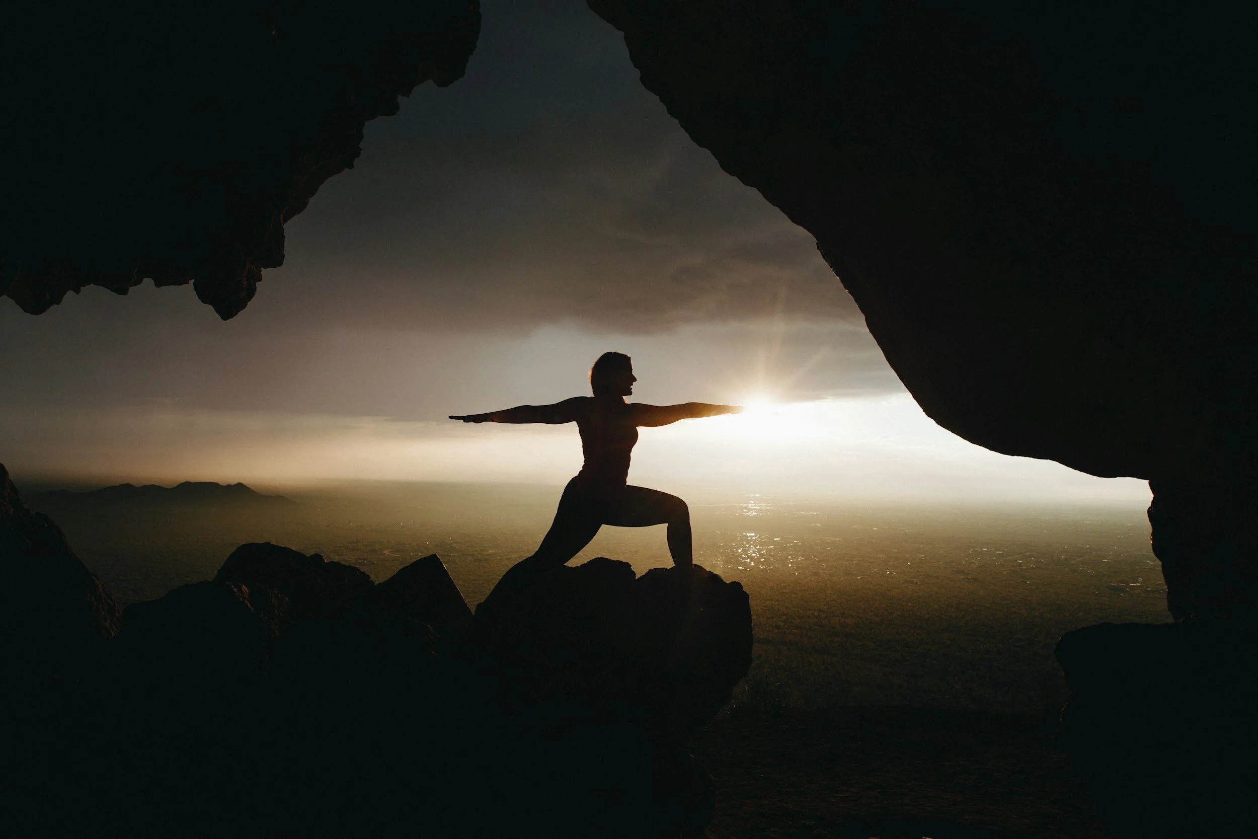 Person practicing yoga in a balancing pose inside a rock formation at sunset with a scenic view of the ocean in the background.