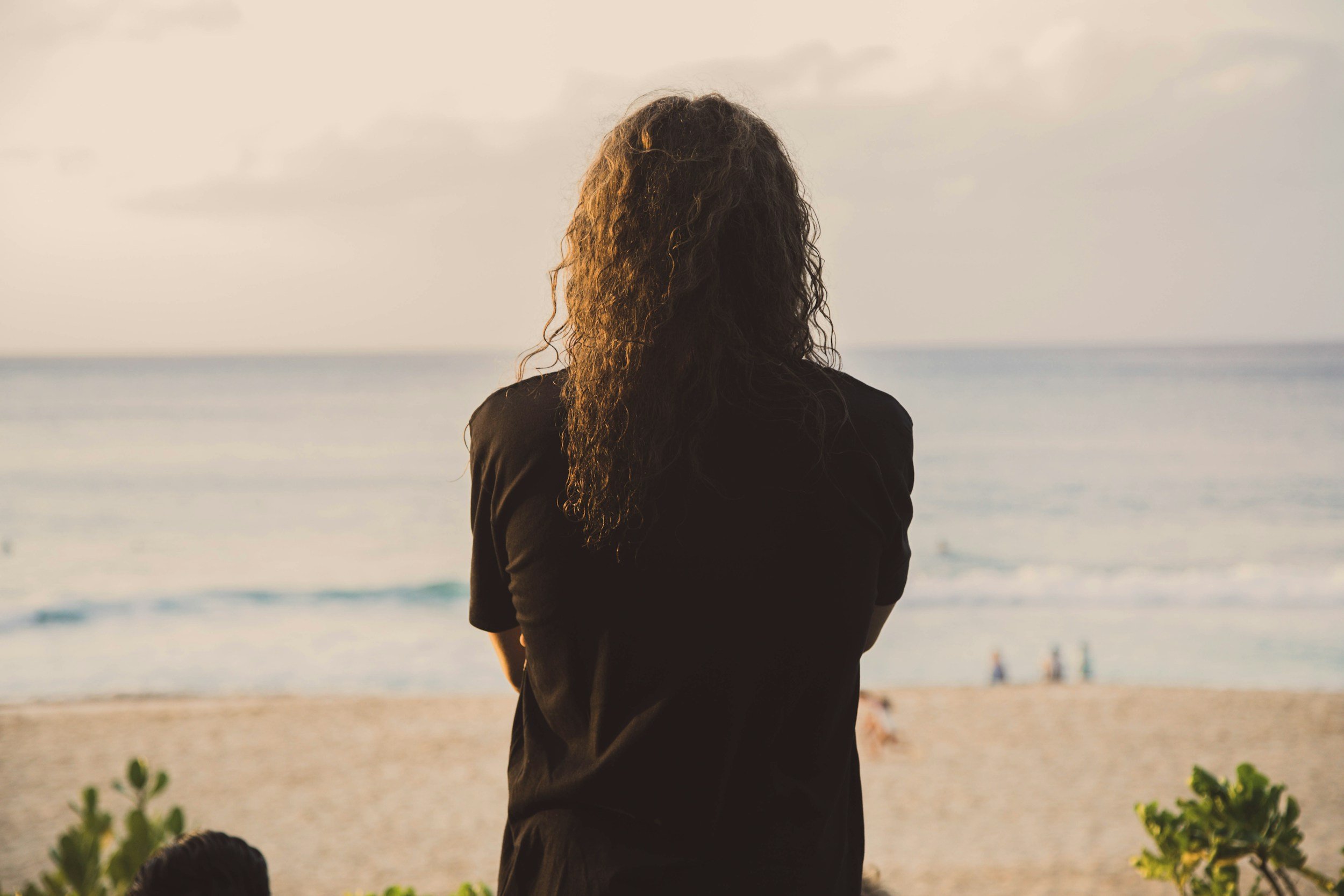 A woman with curly hair standing on a beach, gazing at the ocean during sunset, with a few people visible in the distance.