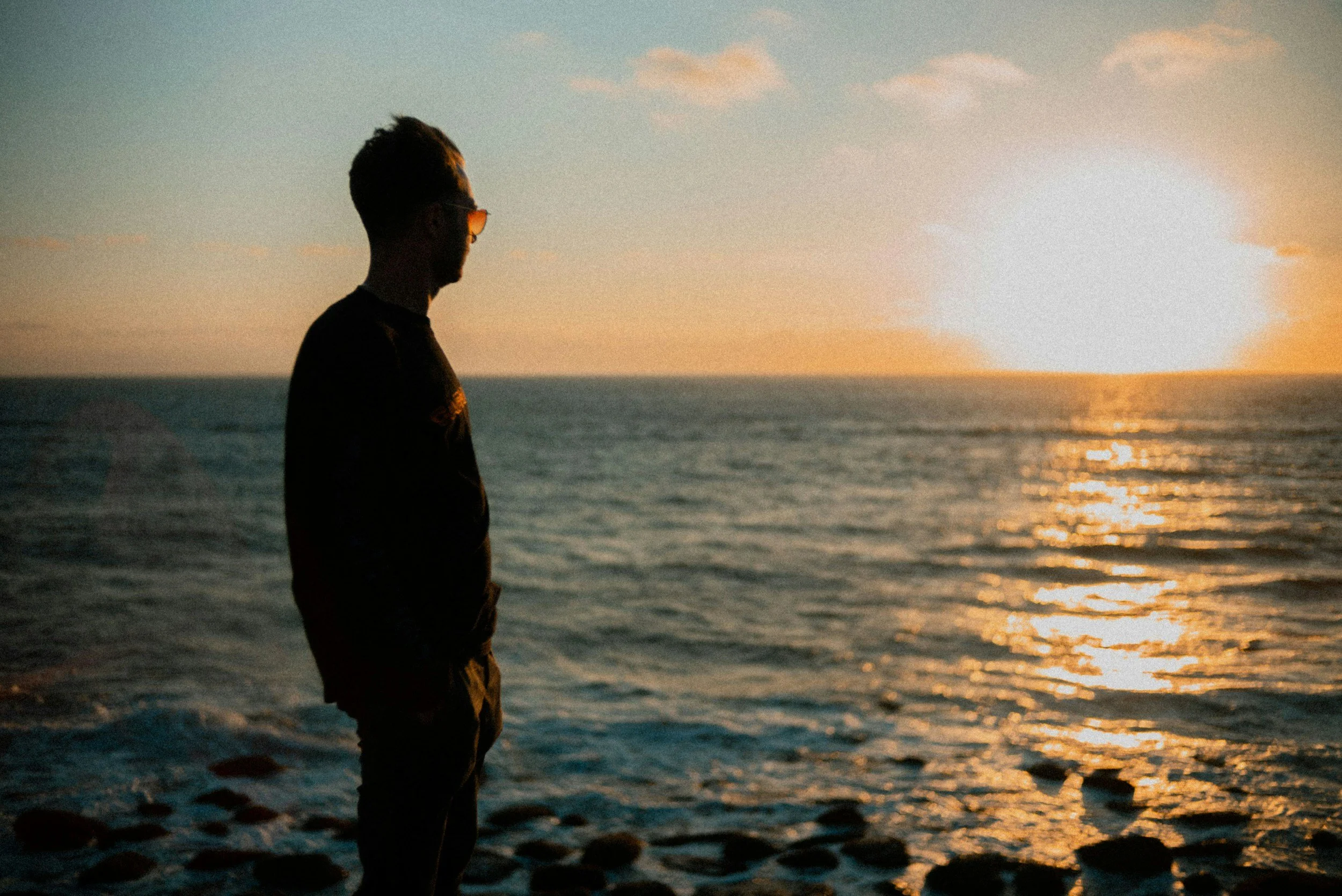 A silhouette of a man with glasses standing on the beach during sunset, gazing at the ocean and the sky with soft clouds.
