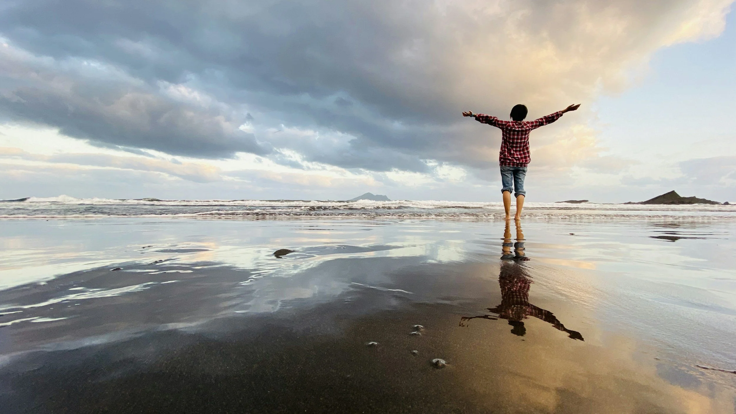 Person stands with arms outstretched on a beach at sunset, reflected in the wet sand, with cloudy sky and distant islands in the background.