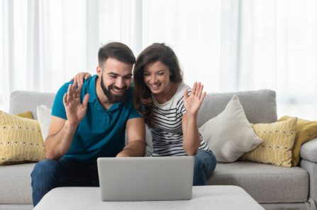 A man and woman sitting on a gray sofa, waving at a laptop, smiling, with yellow and beige pillows behind them in a bright living room.