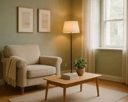 Living room with beige sofa, wooden coffee table, floor lamp, green plant, and framed pictures on the wall near a window with curtains.