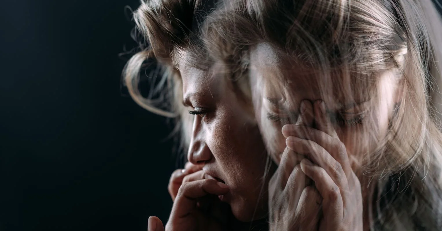 Close-up of a woman with blonde hair, appearing deep in thought or emotional distress, with hands clasped near her face and eyes closed.