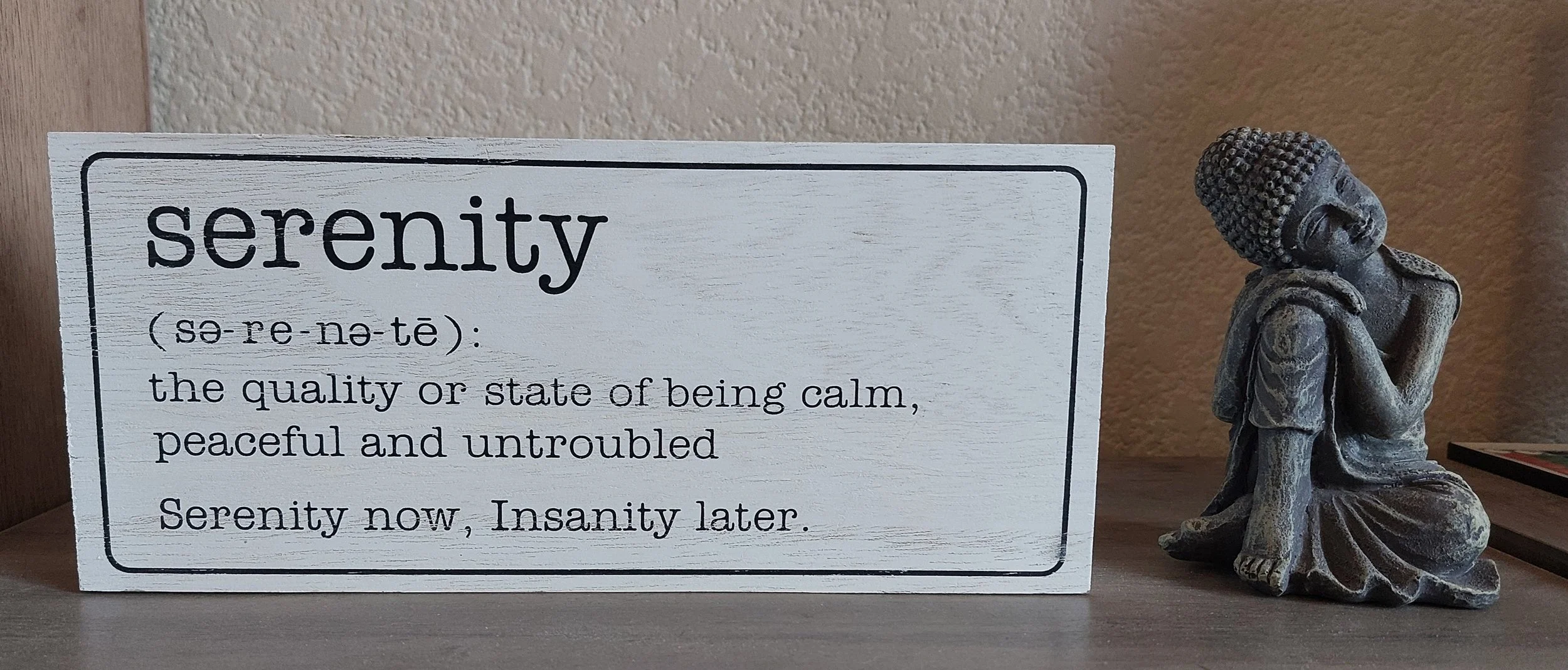 A decorative wooden sign with the word 'serenity' and its definition, placed next to a statue of a meditating Buddha.