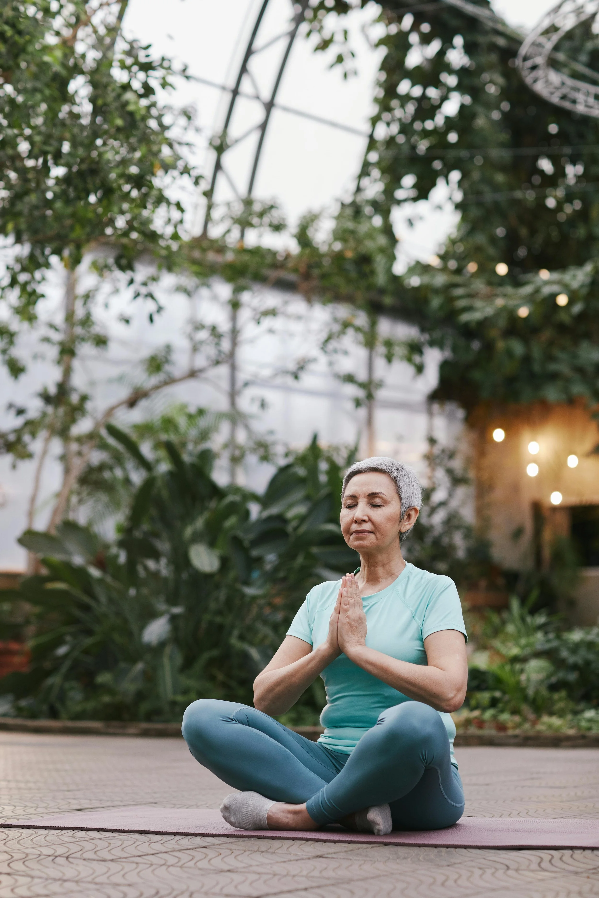 A woman practicing yoga outdoors in a meditative pose surrounded by greenery, sitting cross-legged on a yoga mat with hands pressed together in prayer position. She appears calm and focused with a serene expression.