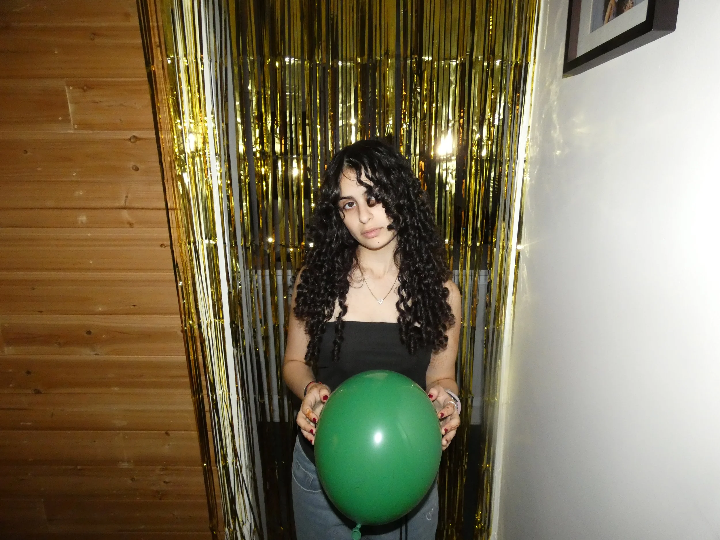 The artist, Libby Ember, standing in front of a striped curtain of gold metallic strips, holding a green balloon.