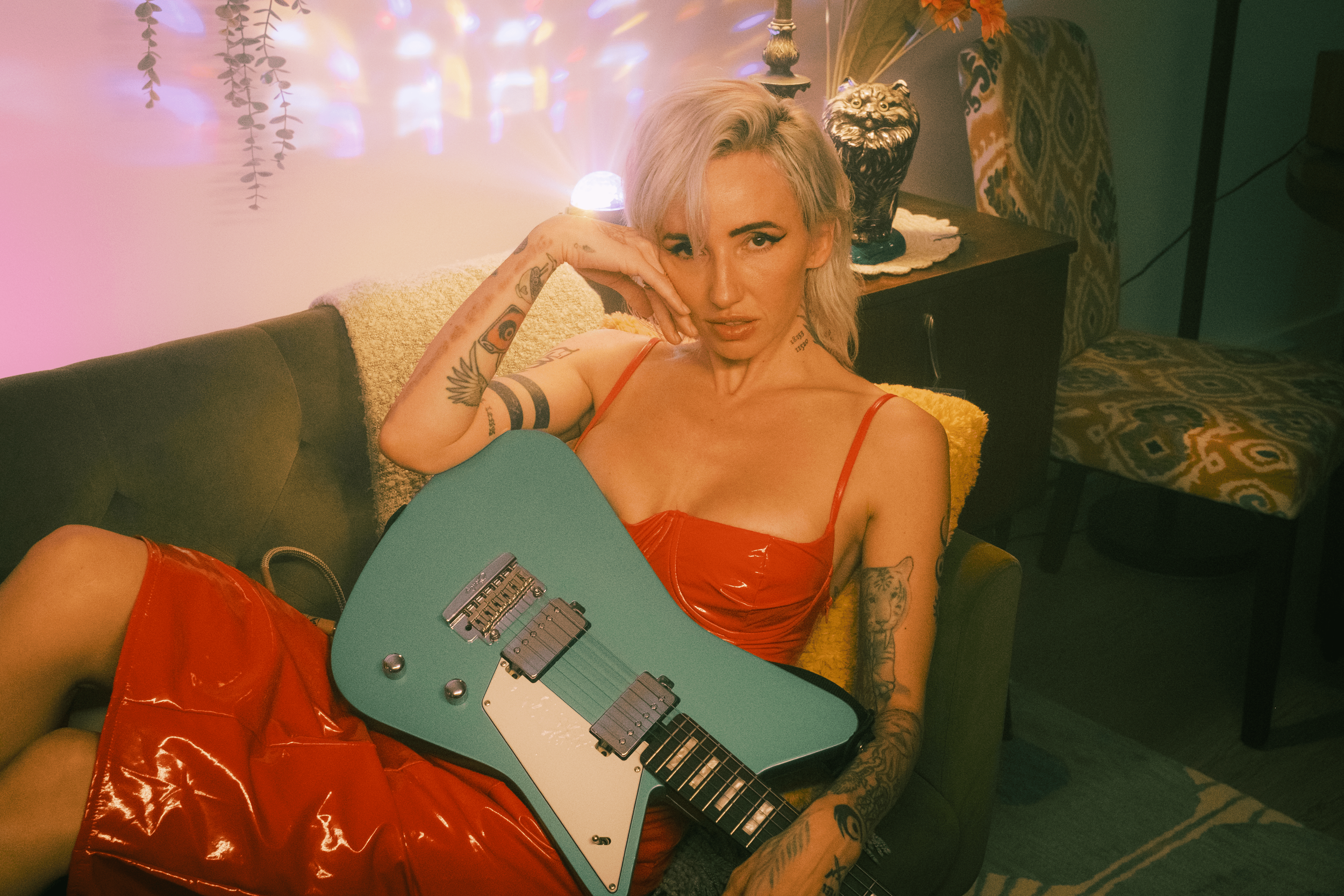 The Artist, Lights, wearing a red dress, sitting on a yellow furry cushion on a sofa, holding a teal electric guitar in her lap, in a dimly lit room with colorful lights and decorative items in the background. Photo Credit- Warwick Hughes.