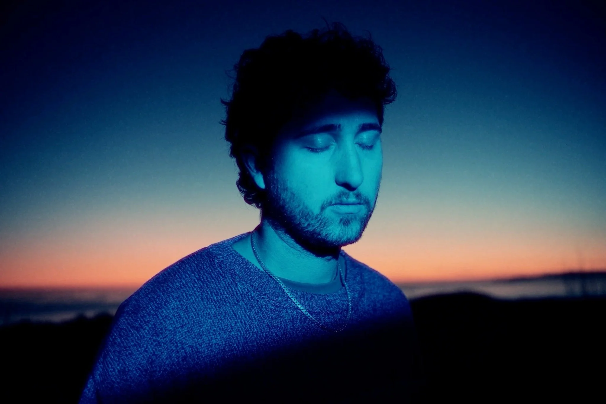 The artist ARK IDENTITY, wearing a sweater, standing outdoors during sunset with a colorful sky in the background.