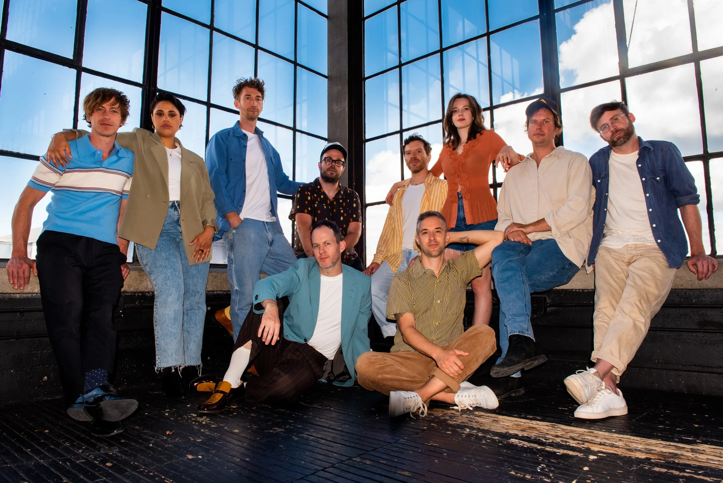 The band, Dwayne Gretzky, posing together indoors in front of large industrial-style windows with a view of blue sky and clouds. They are dressed casually, some standing and some sitting.