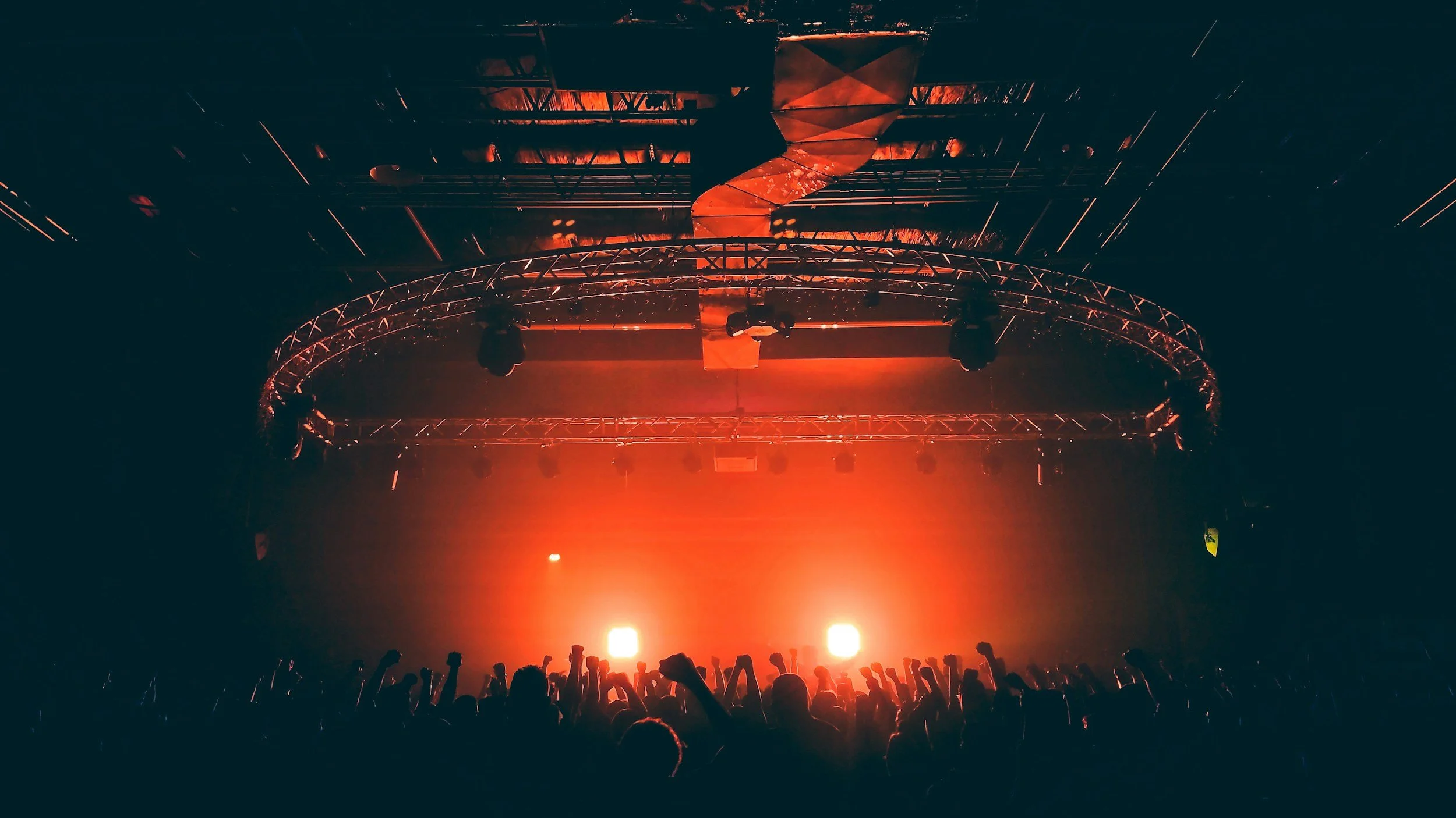 A concert stage with red lighting, a crowd with raised hands, and a rigging structure overhead.