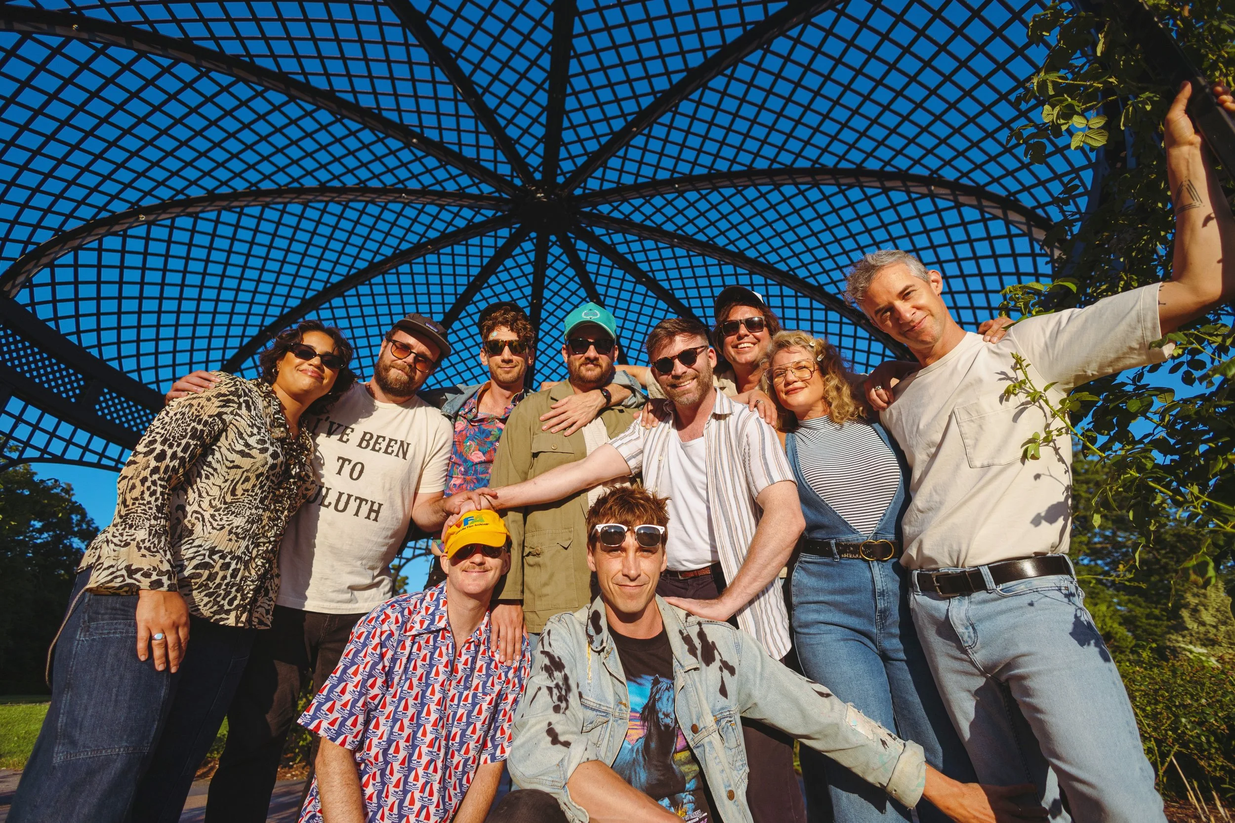 Dwayne Gretzky posing for a photo under a large, open, lattice metal structure on a sunny day, with some greenery visible.