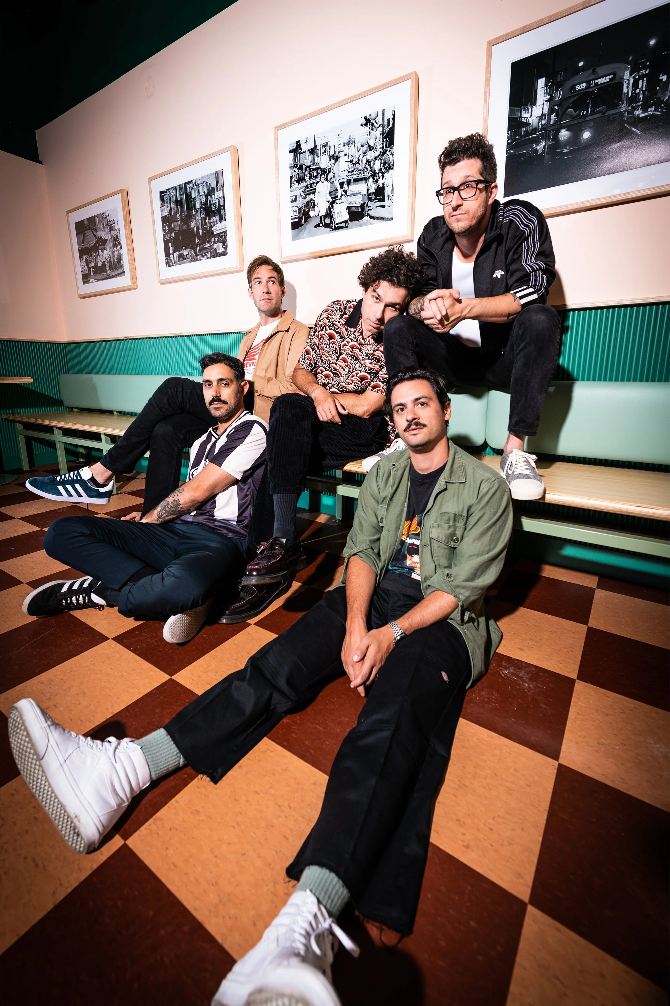 The band, Arkells, sitting and posing in a room with framed black and white photos of city scenes on the wall, wearing casual clothing. Photo by Nathan Nash.