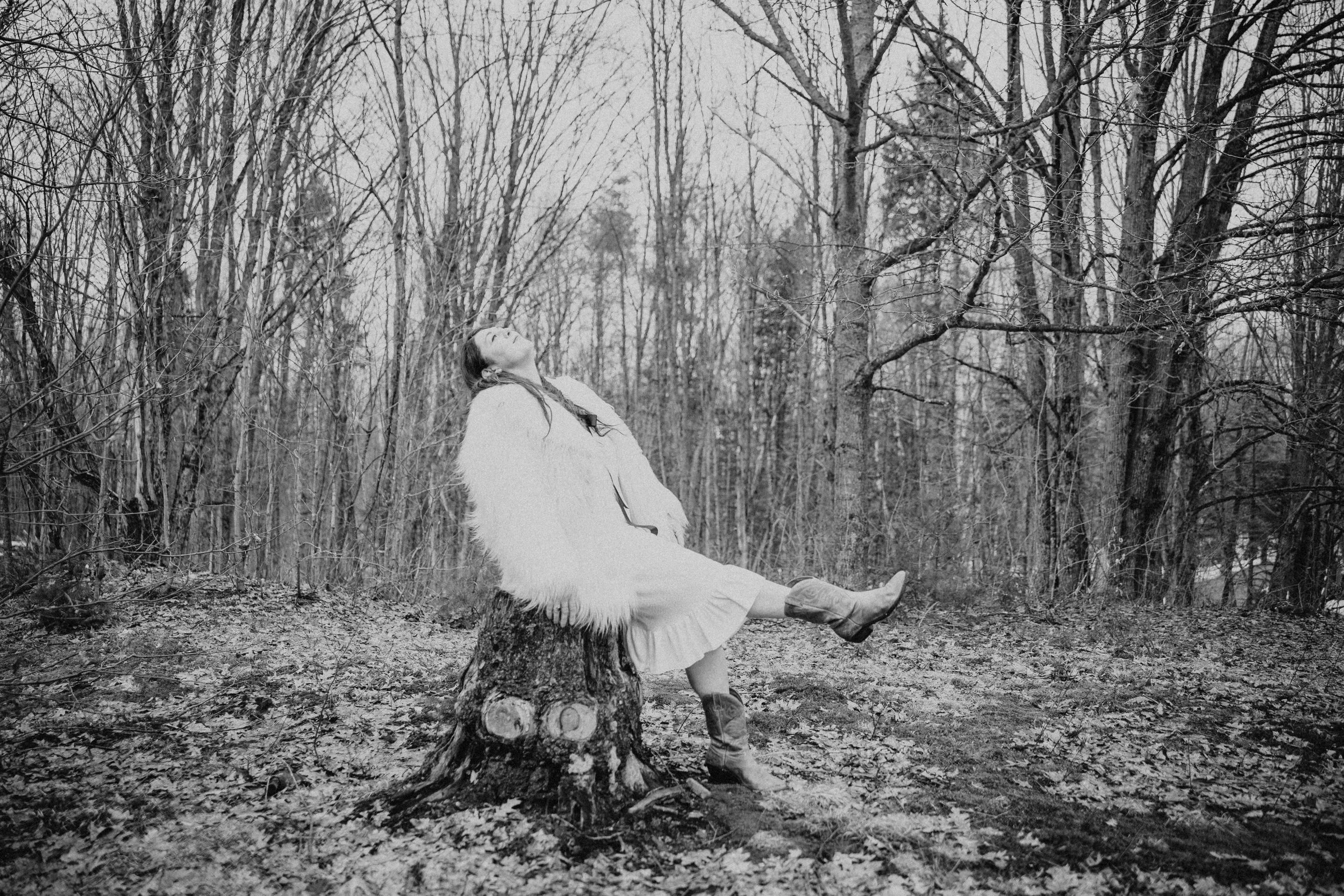 Artist, Terra Lightfoot, sits on a tree stump in a wooded area, looking upward with her eyes closed.