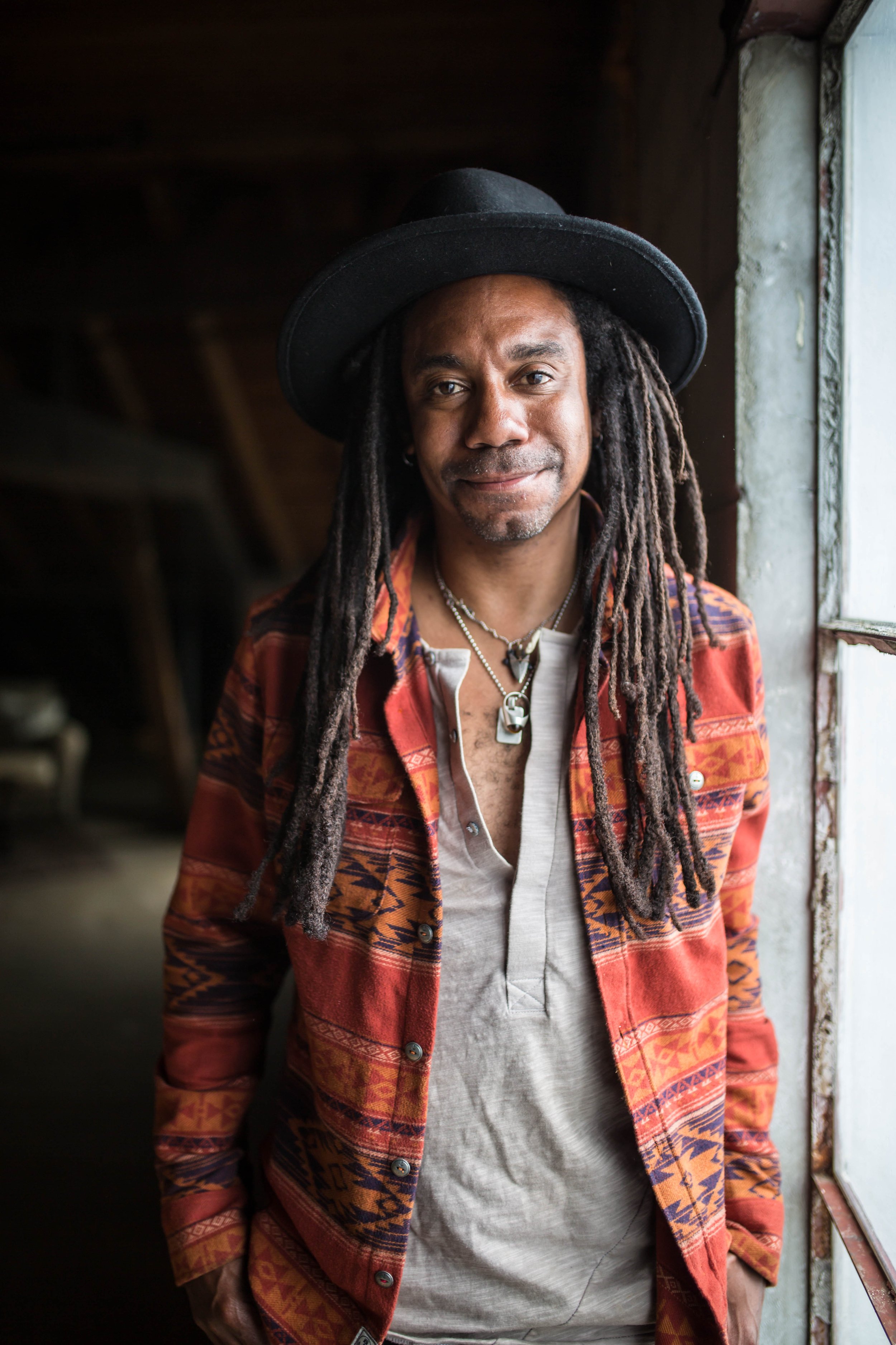 The artist, Julian Taylor, wearing a black hat, colorful patterned jacket, and necklaces, standing near a window in a dimly lit space.