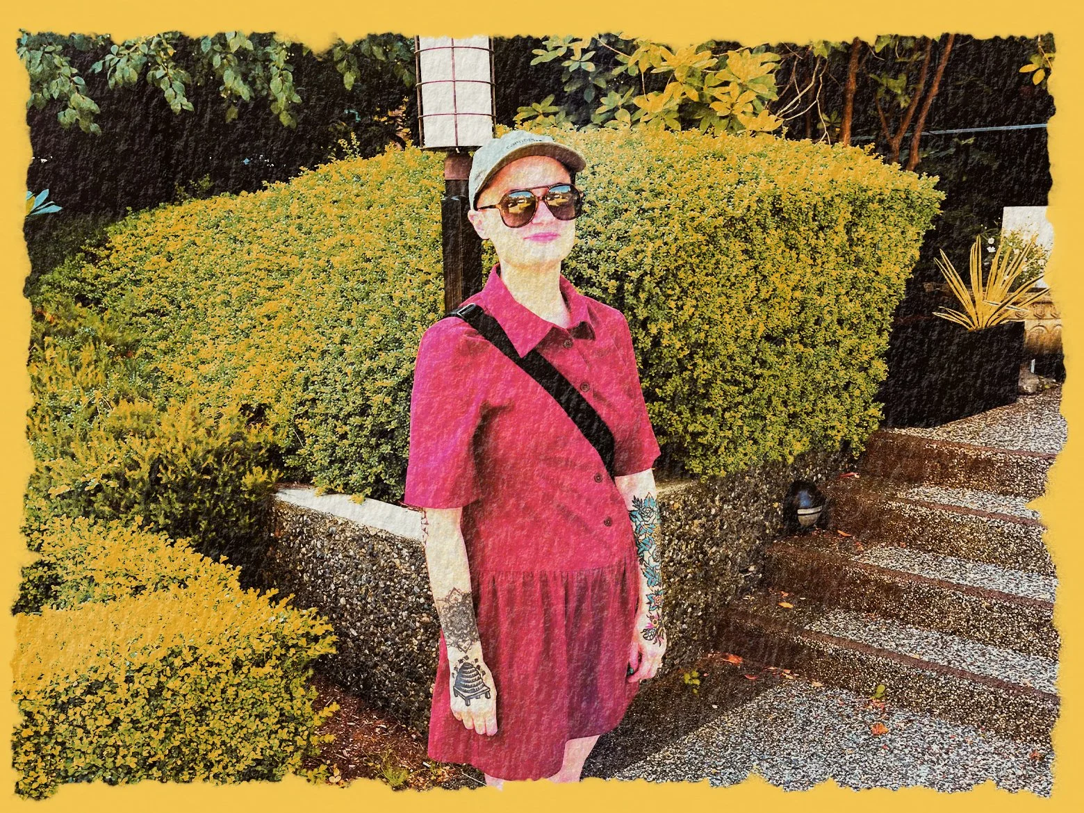 A woman with tattoos on her arms and floral tattoos on her hands stands outdoors next to a bush and steps, wearing a pink dress, sunglasses, and a gray cap, with a black crossbody bag.
