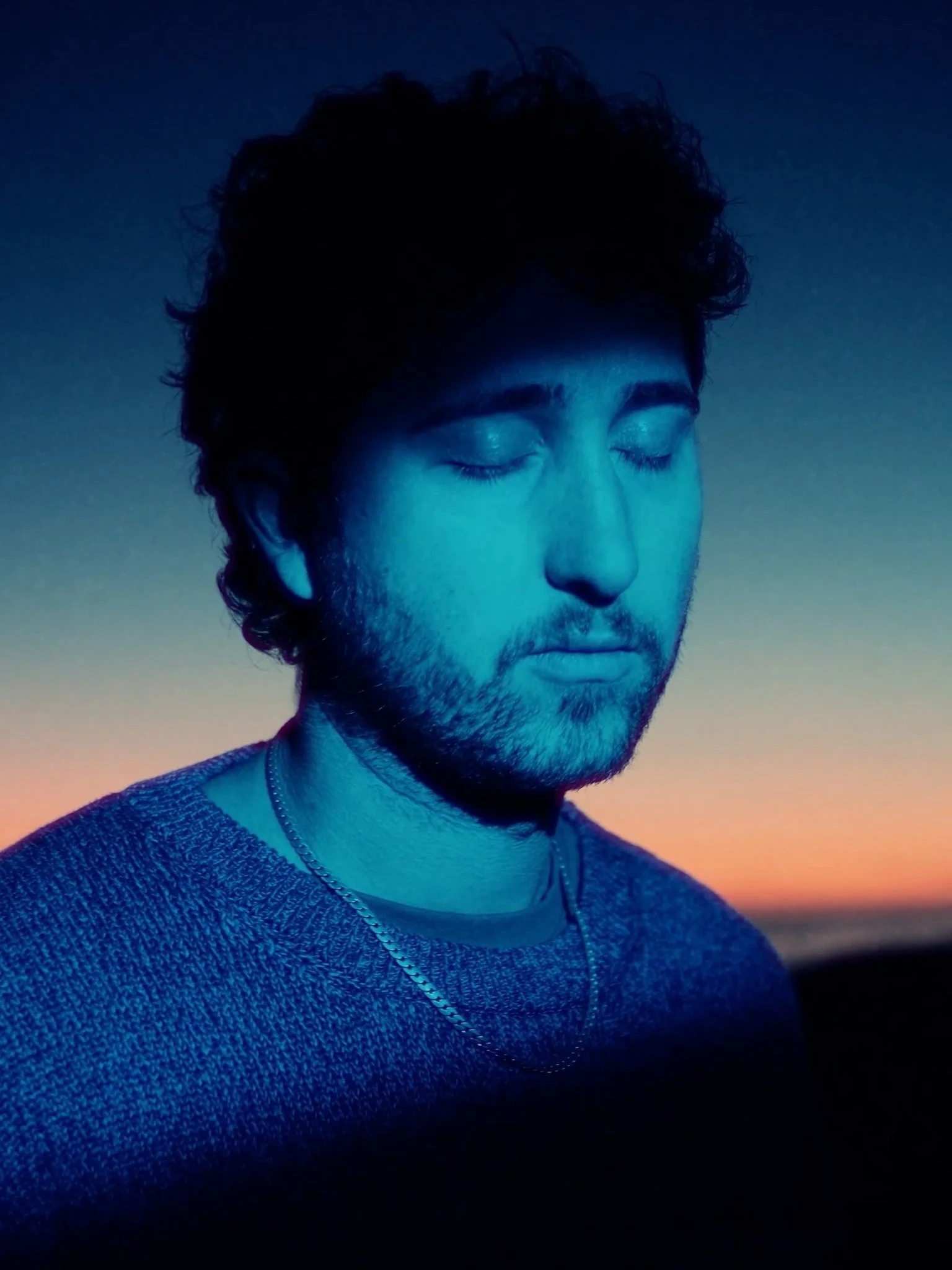 The artist, ARK IDENTITY, with his eyes closed. He is wearing a knit sweater and a silver necklace. The background features a sunset or sunrise sky with a gradient of orange, yellow, and blue hues.