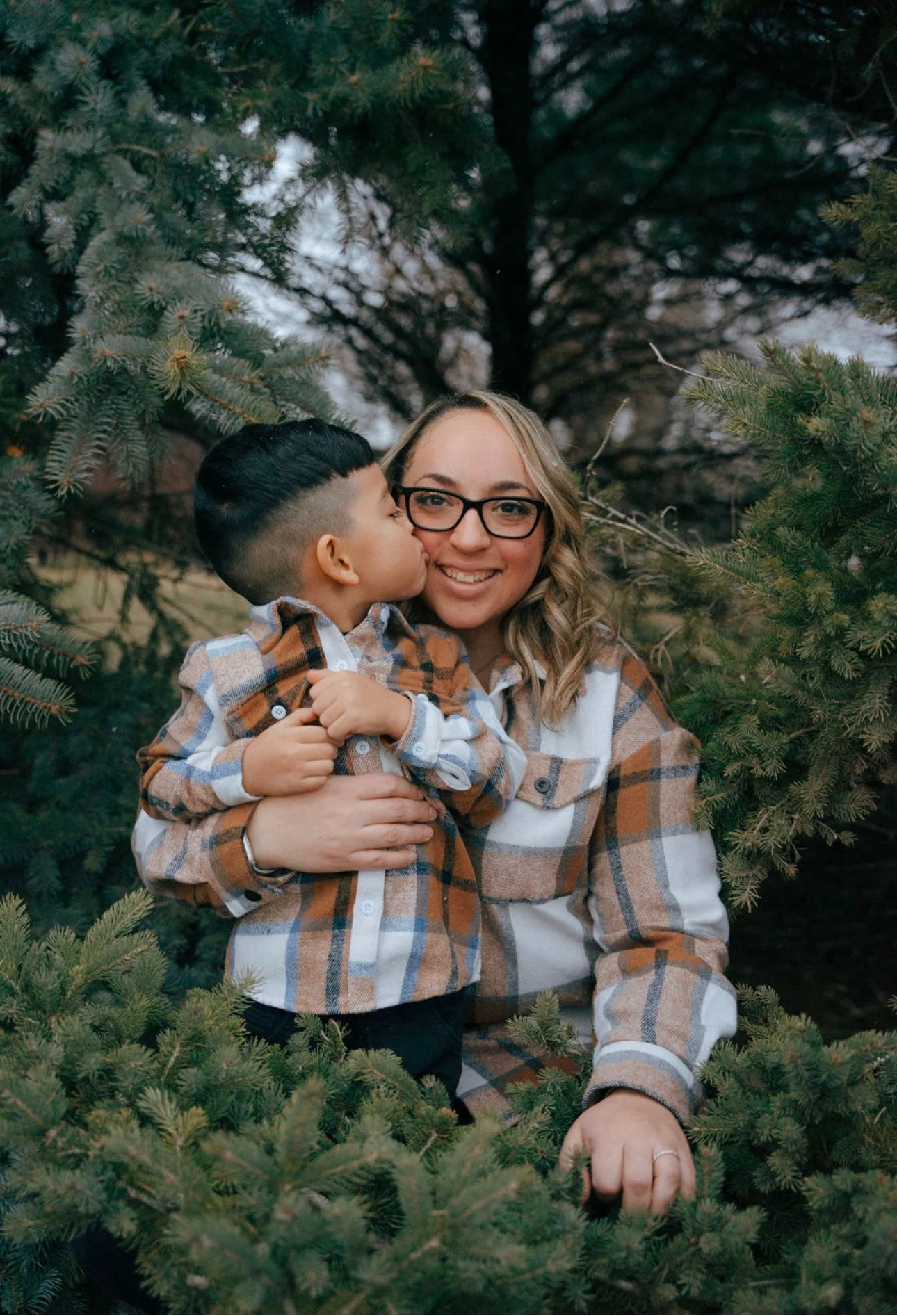 A woman with glasses and blonde hair smiles while holding a young boy with black hair, giving him a kiss on the cheek, surrounded by green pine tree branches.