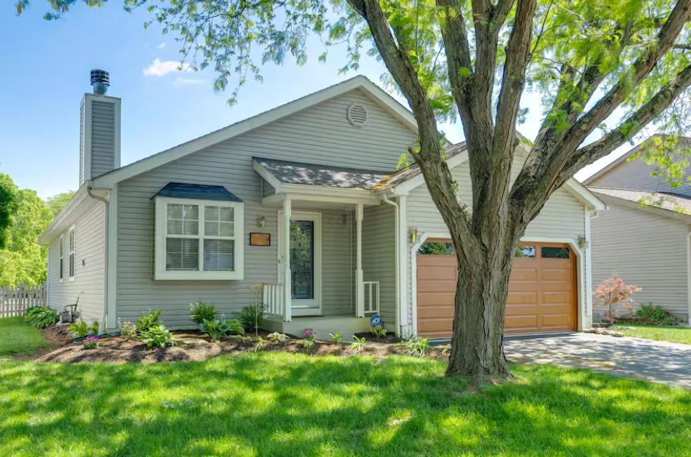 A suburban house with grey siding, a small front porch, a front yard with green grass, a large tree, and a garage with a brown door.