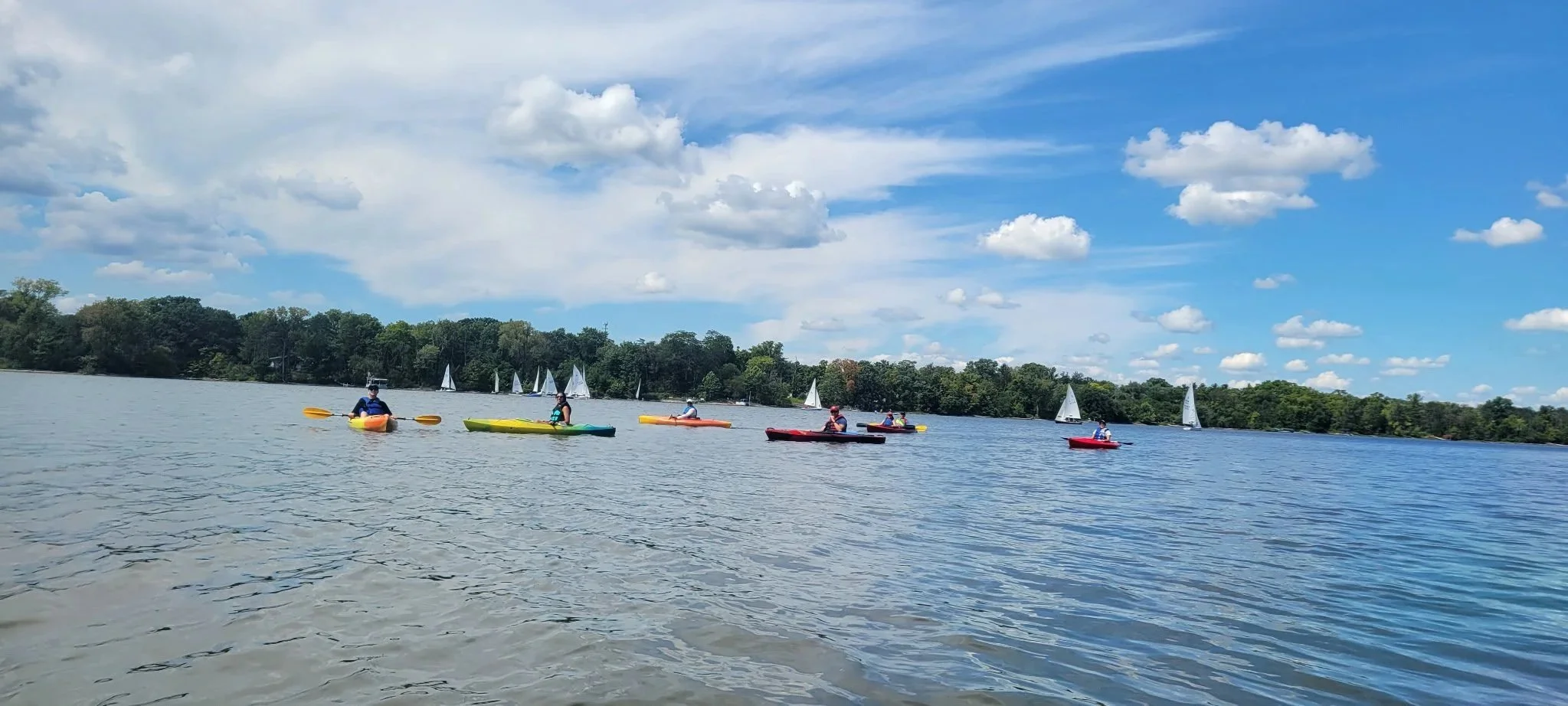 People kayaking on a lake with sailboats in the background under a partly cloudy sky.