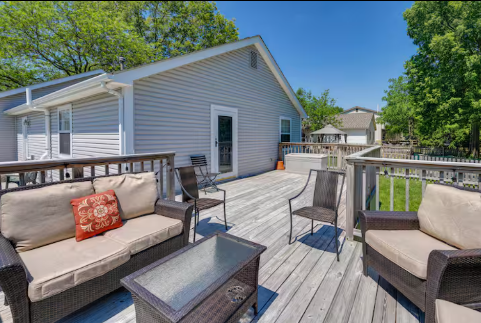 Outdoor wooden deck with wicker furniture, including a sofa with beige cushions and a red decorative pillow, and several chairs, attached to a house with vinyl siding, surrounded by green trees and a blue sky.